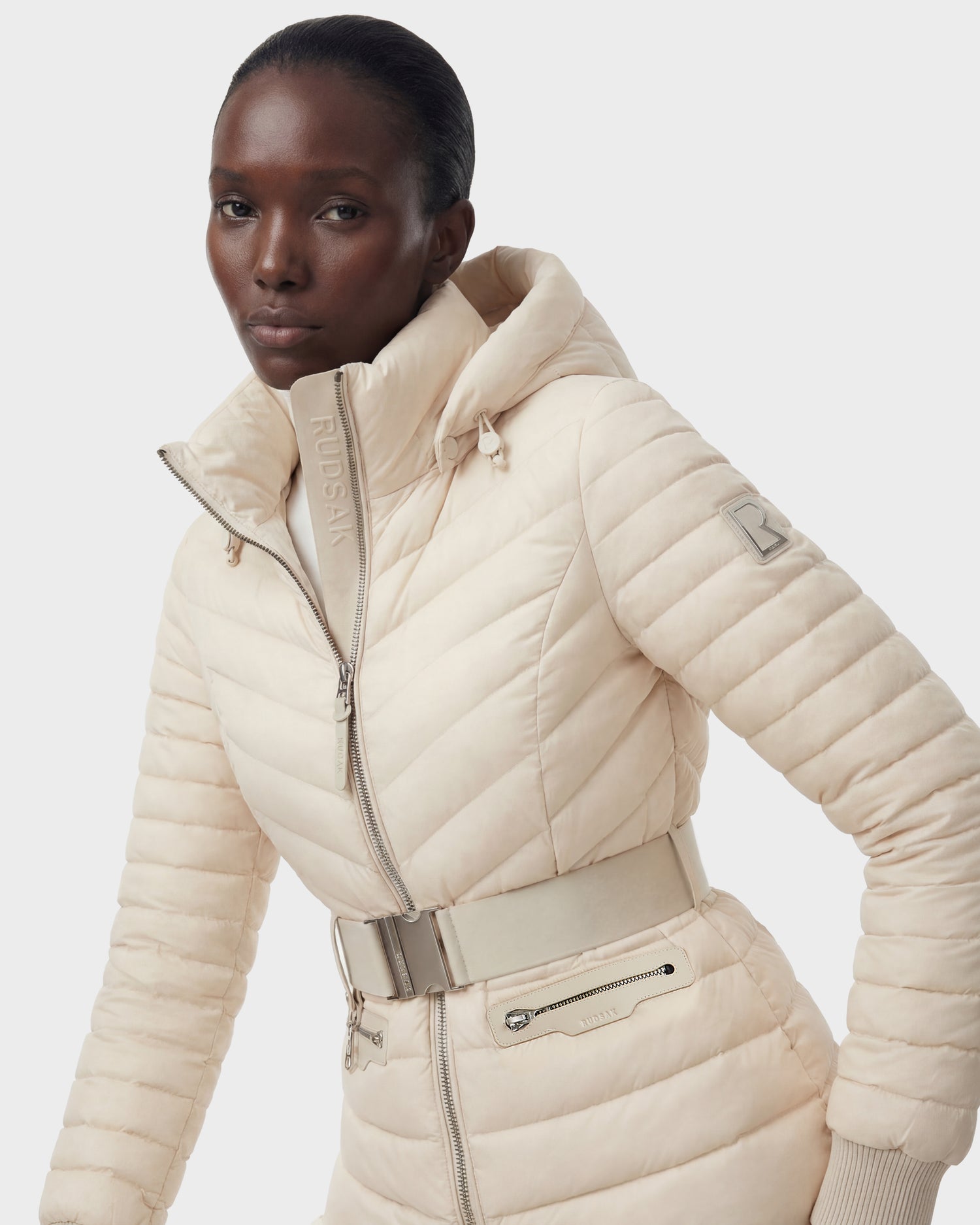 A person wears the Rudsak SCARLETT - 8125004 Vanilla women's lightweight down jacket with hood, vegan leather waist belt, silver zippers, and RUDSAK branding on the collar and sleeve. The background is plain white.