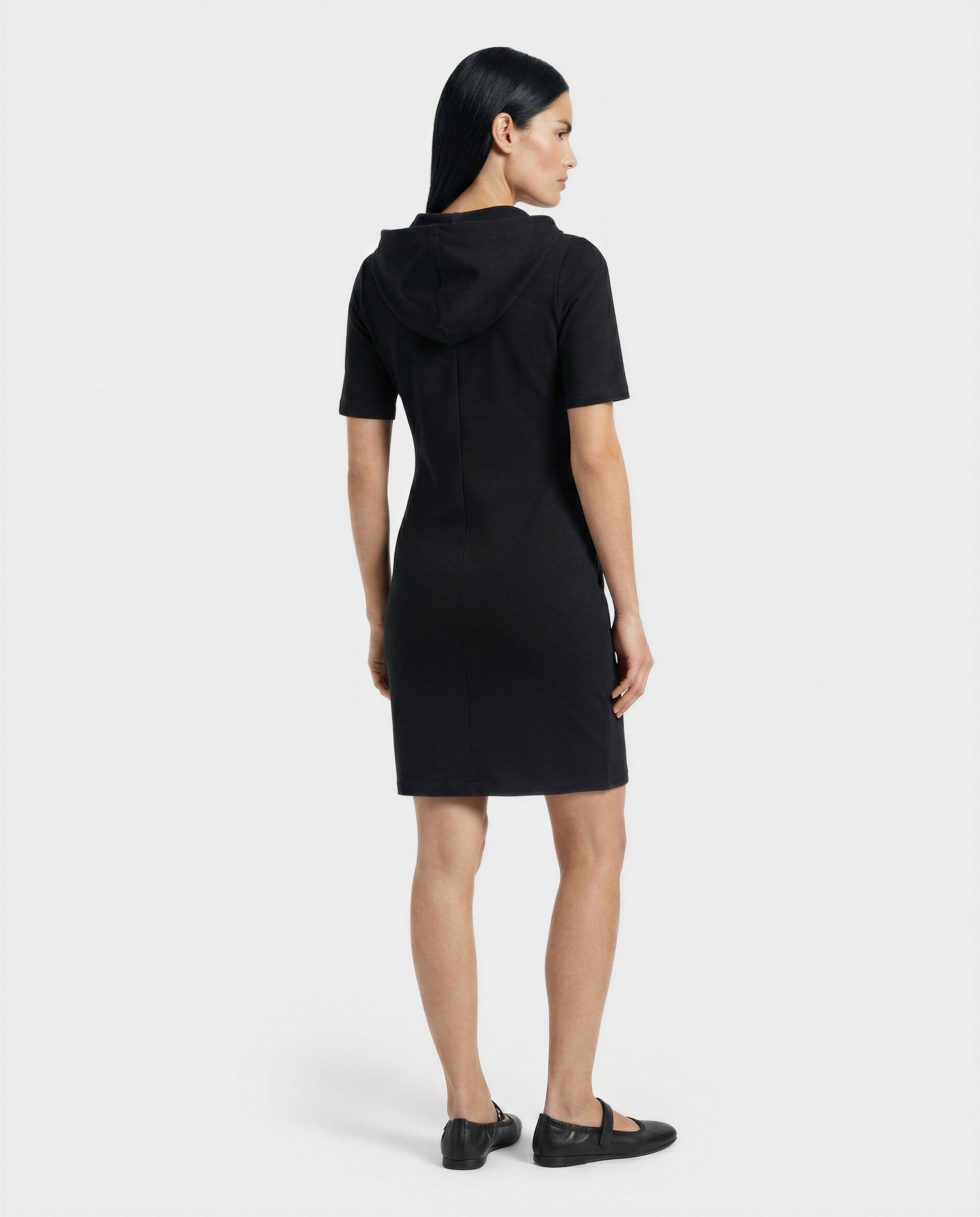 A woman with straight black hair faces away in a knee-length, short-sleeved, hooded black dress—GISELA - L524004 Black by Rudsak—with black flat shoes, set against a plain white background.