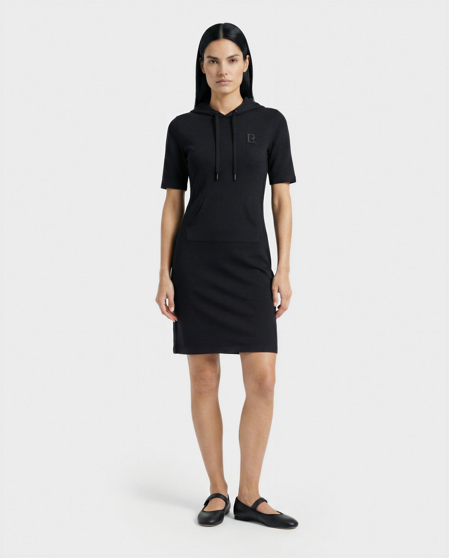 A woman with straight dark hair models the Rudsak GISELA - L524004 Black short-sleeve hooded dress with a front pocket and black flats, standing against a plain white background.