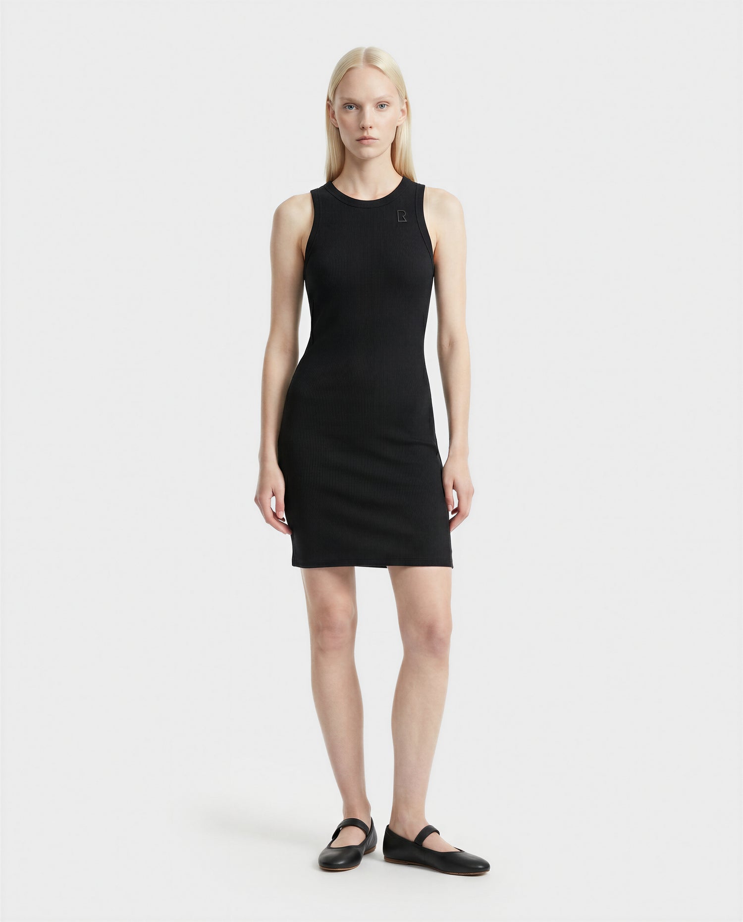 A woman with straight, light blonde hair stands against a white background, wearing the Rudsak ADELLA - L524005 Black sleeveless fitted dress and black flat shoes, facing forward with a neutral expression and arms at her sides.