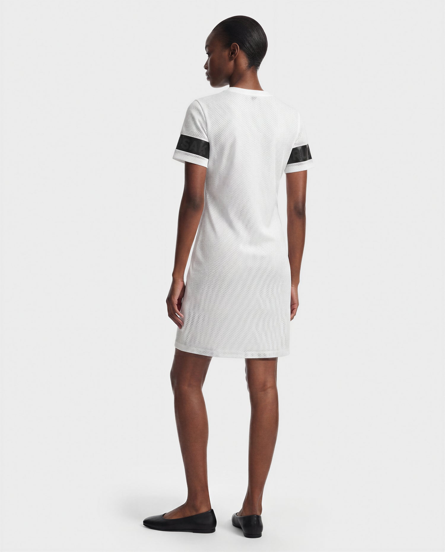 A person stands facing away, wearing the Rudsak ELAINE - L523029 Optic White short-sleeved dress with black striped sleeves and black flat shoes, against a plain light background.