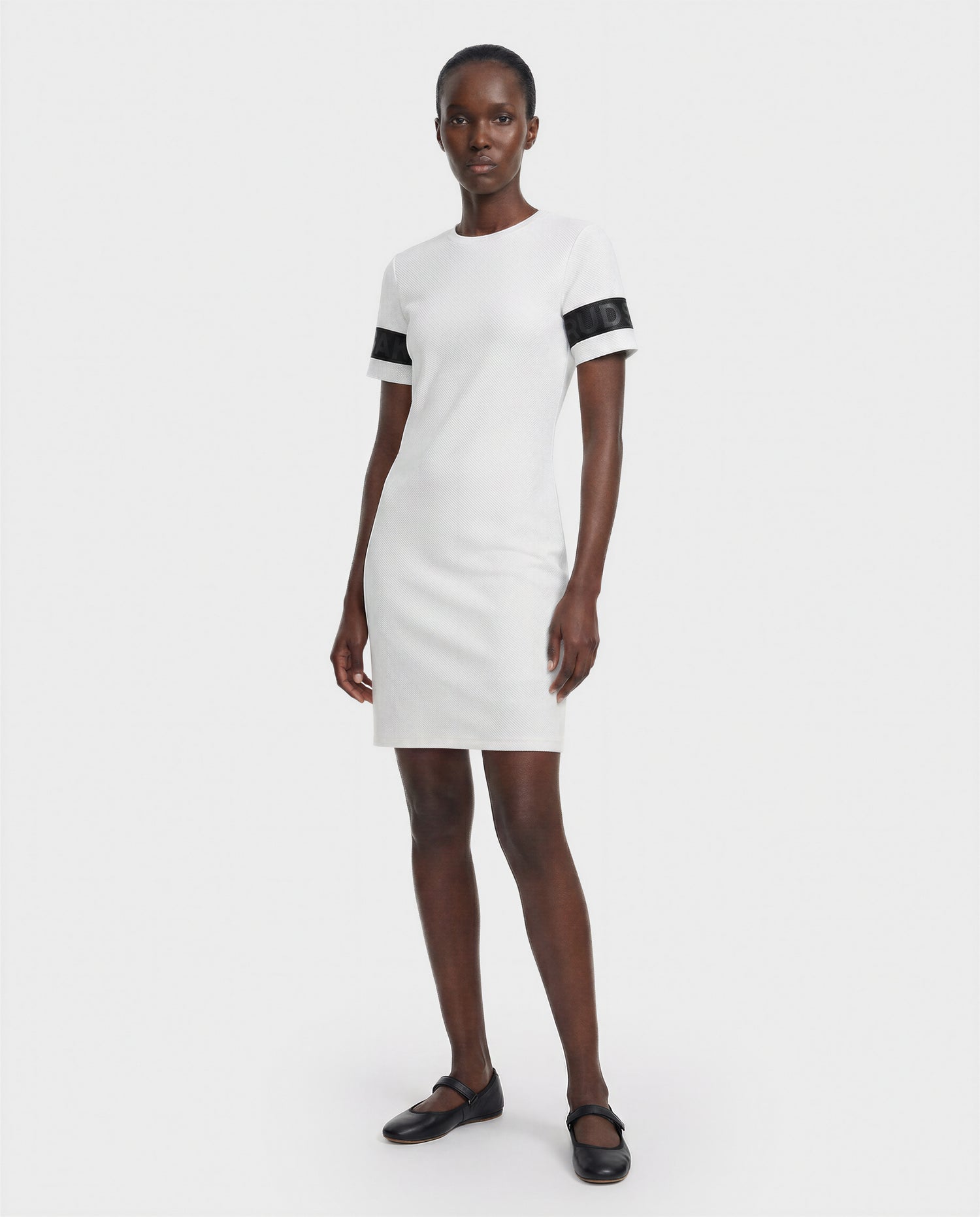 A woman with short hair wears the Rudsak ELAINE - L523029 Optic White dress, featuring short sleeves and black sleeve bands, paired with black flats against a plain white background.