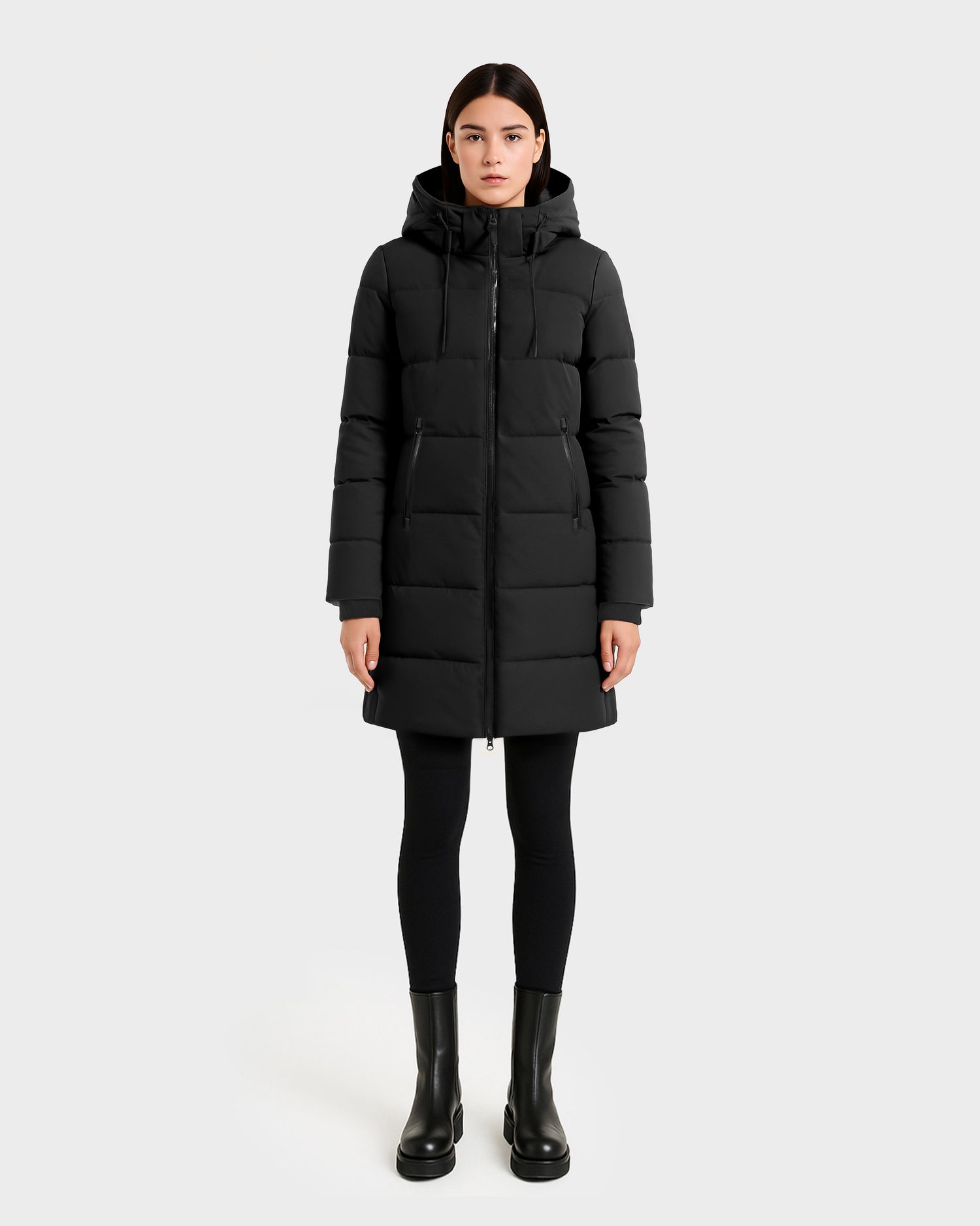 A woman faces forward in a Rudsak SYDNEY COM - 4123511 Black wind-resistant puffer coat with a hood, paired with black leggings and boots, set against a plain light gray background.