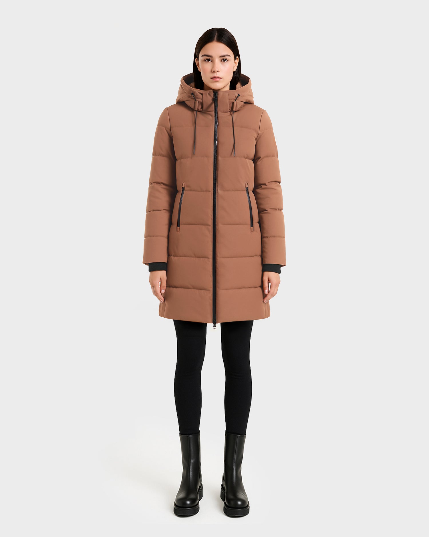 A woman faces forward wearing the Rudsak SYDNEY COM - 4123511 Taupe, a long, water-repellent and wind-resistant duck down parka, paired with black leggings and boots against a plain white background.