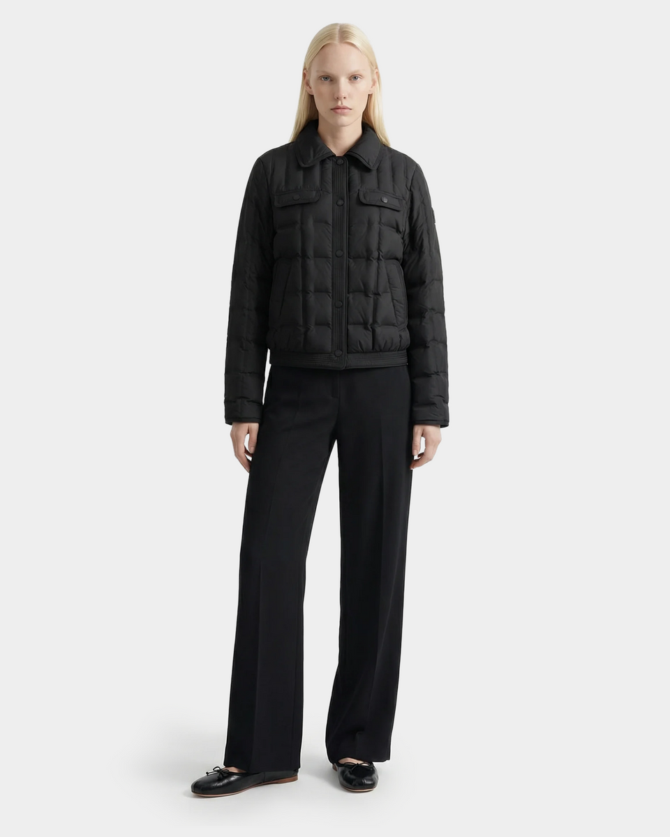 A person with long blonde hair wears the Rudsak SEOUL - 8126027 Black quilted down-fill shirt jacket, black wide-leg trousers, and black loafers, standing against a plain light background for lightweight warmth.