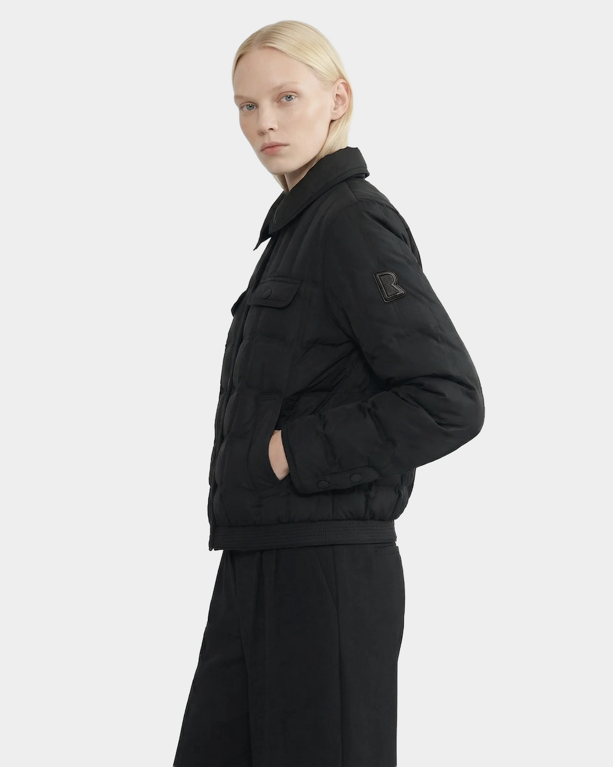 A person with straight blonde hair models Rudsak's SEOUL - 8126027 Black quilted down-fill shirt jacket and black pants, standing sideways against a light background. Lightweight warmth is provided by RDS-certified duck down.