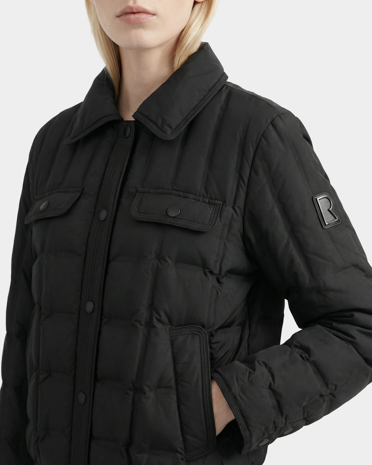 A person with light skin and blonde hair, one hand in the pocket, wears the Rudsak SEOUL - 8126027 Black quilted down-fill shirt jacket featuring flap chest pockets, a collar, and an "R" patch on the left sleeve—perfect for lightweight warmth on chilly days.