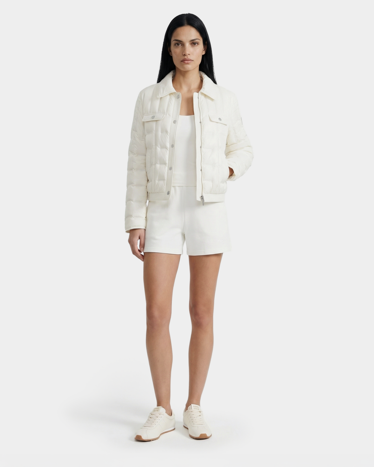 A woman with long dark hair models the Rudsak SEOUL - 8126027 Iceland white quilted down-fill shirt jacket, tank top, shorts, and sneakers—standing against a white background and showcasing effortless lightweight warmth.