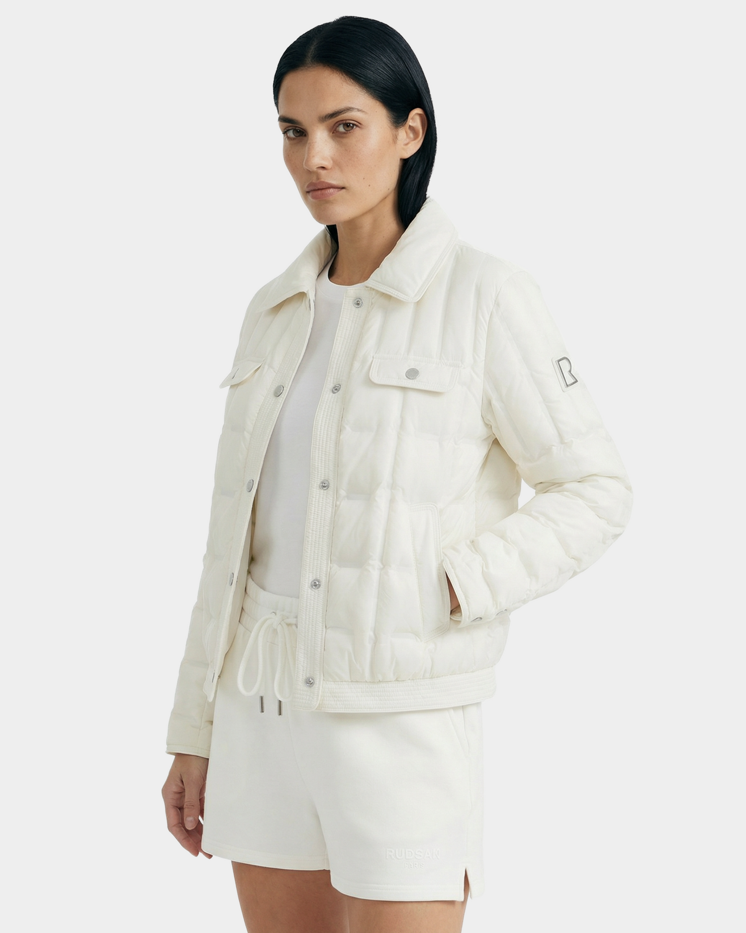 A woman with straight black hair models the Rudsak SEOUL - 8126027 Iceland, a white quilted down-fill shirt jacket, over a white shirt and matching shorts against a plain light background for lightweight warmth.