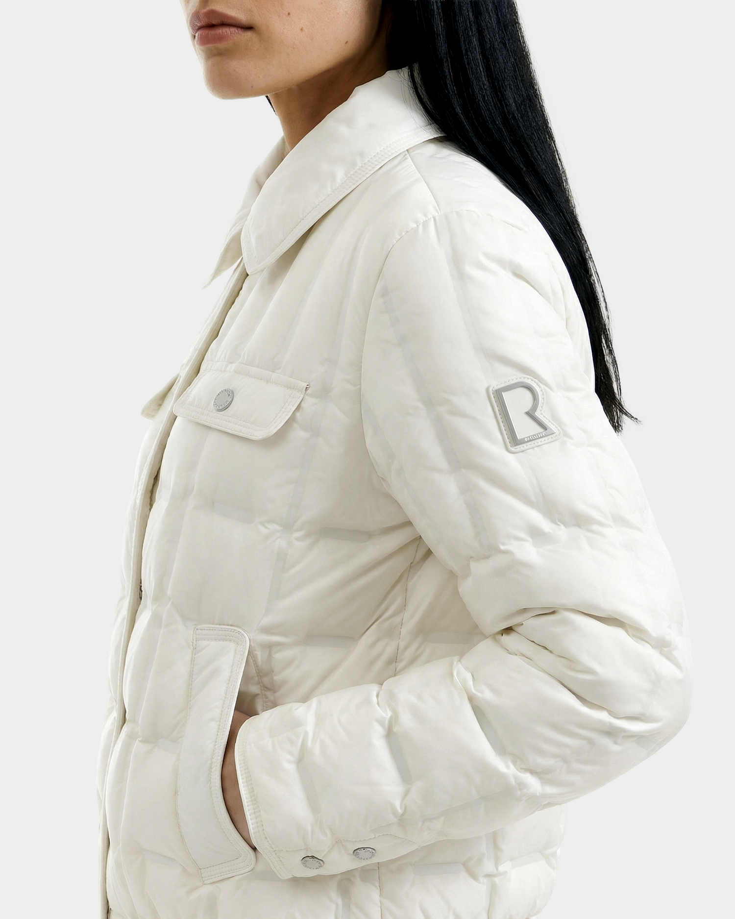 A woman with long dark hair wears the Rudsak SEOUL - 8126027 Iceland light puffy down-fill shirt jacket with flap pockets, hand in one pocket. An embroidered R logo appears on the upper sleeve. The background is plain white.