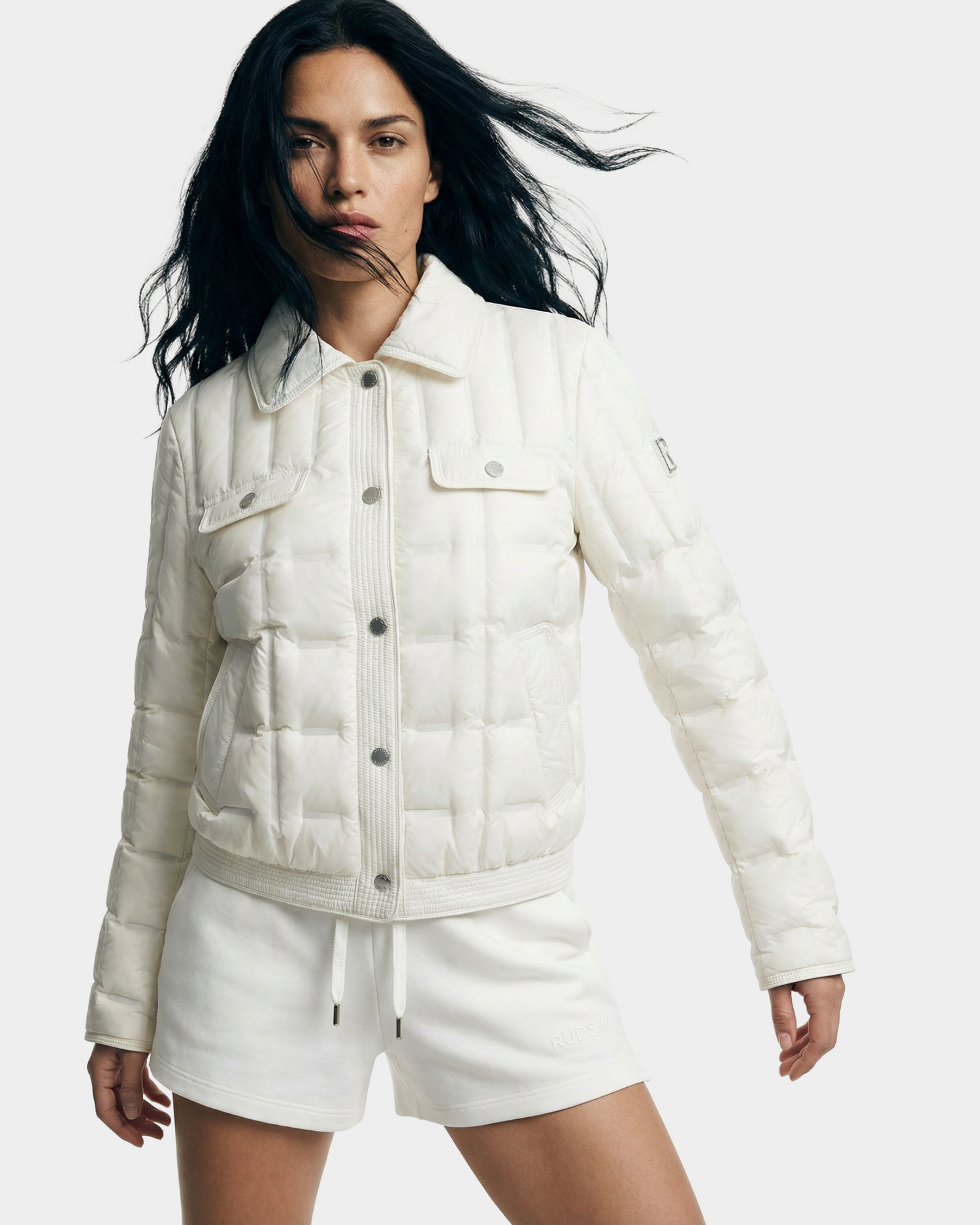 A woman with long dark hair wears the Rudsak SEOUL - 8126027 Iceland white quilted down-fill shirt jacket and white shorts, standing against a plain light background, showcasing its effortless style and lightweight warmth.