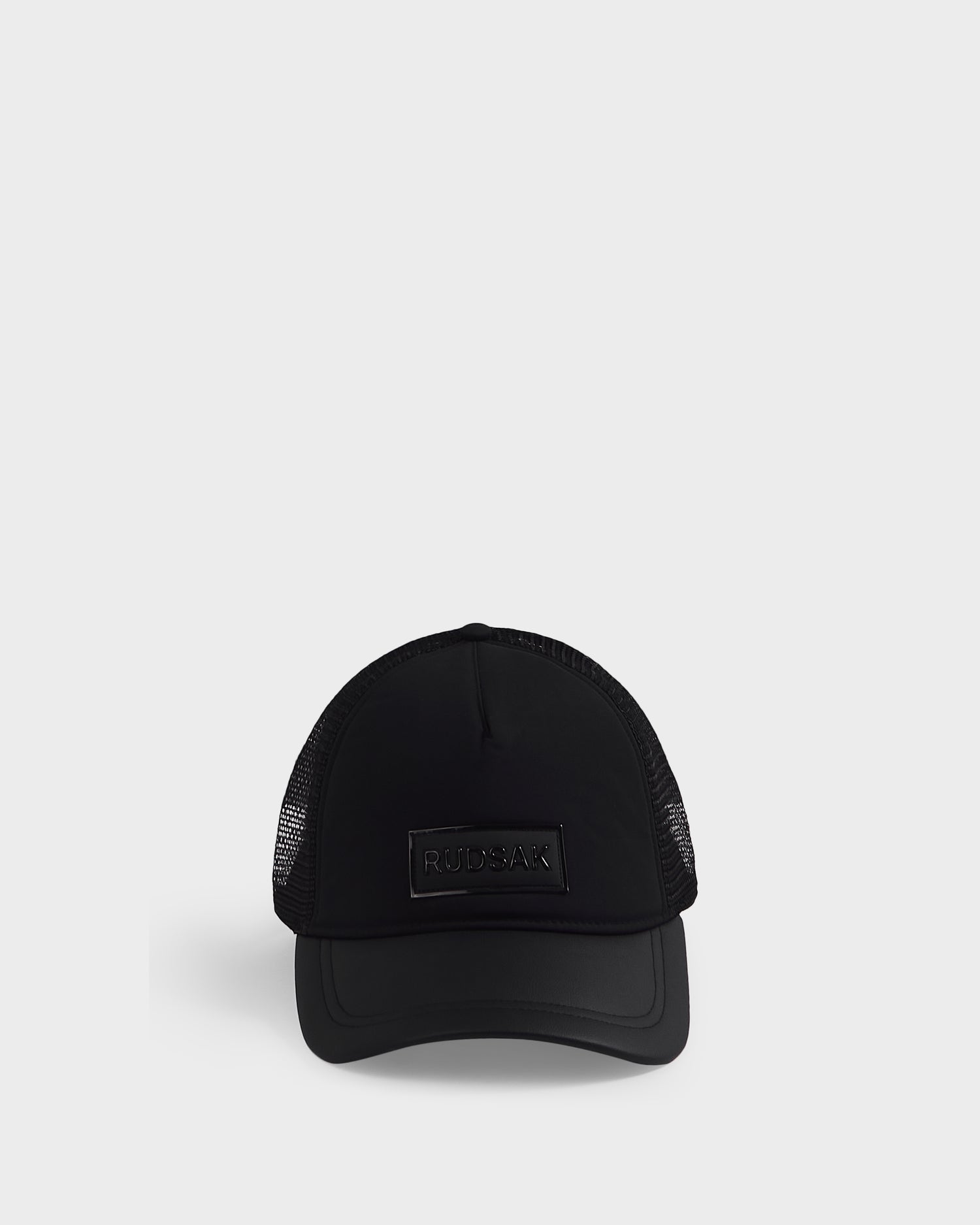 The FENIX - 9422522 Black by Rudsak is a black trucker cap with mesh sides and back, featuring a rectangular Rudsak logo patch on the front, shown against a plain white background.