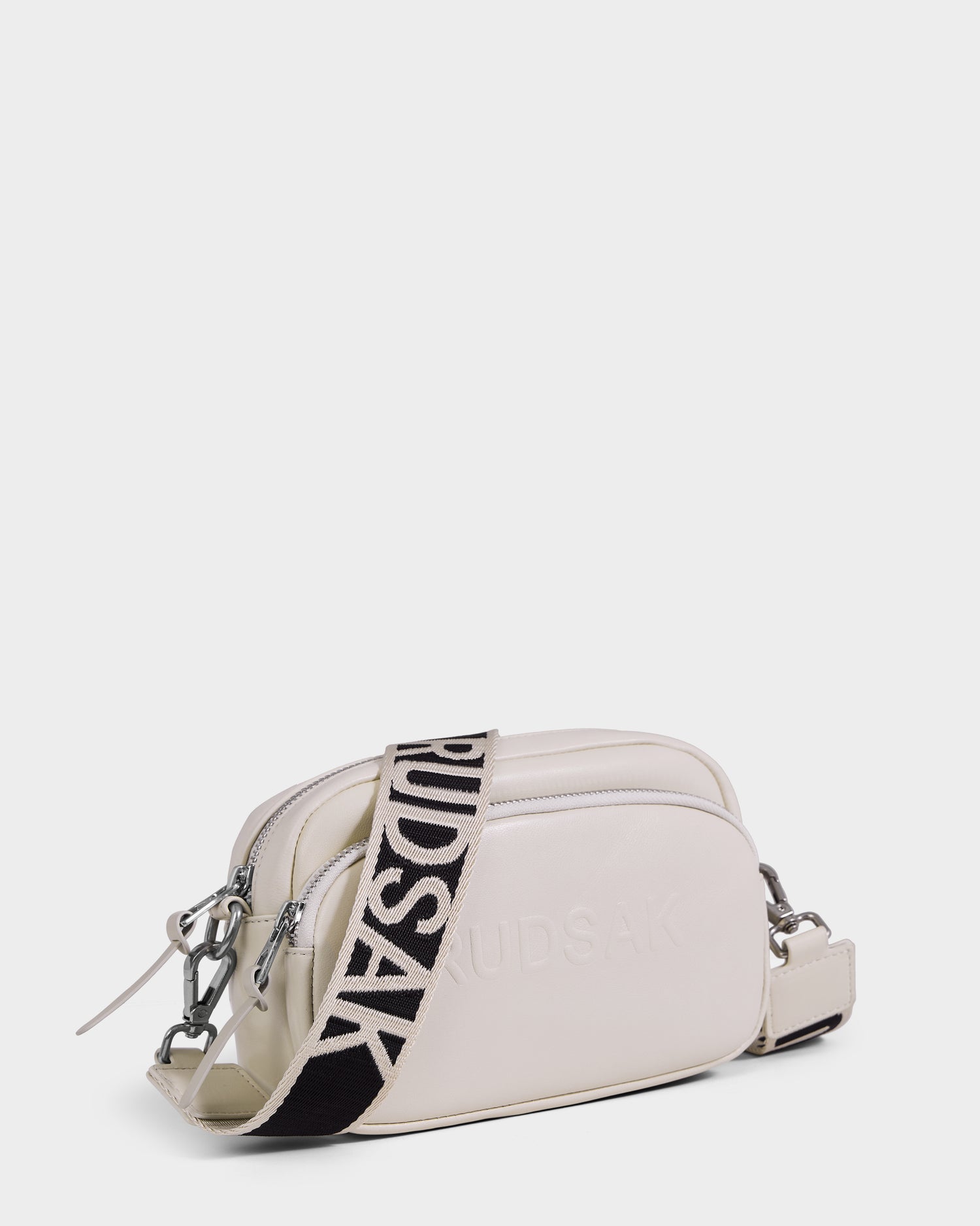 The Rudsak ZOYA L - 8325543 Vanilla is a small ivory leather crossbody bag with two zip compartments, silver hardware, a wide adjustable black strap with bold white RUDSAK lettering, and embossed branding on the front.