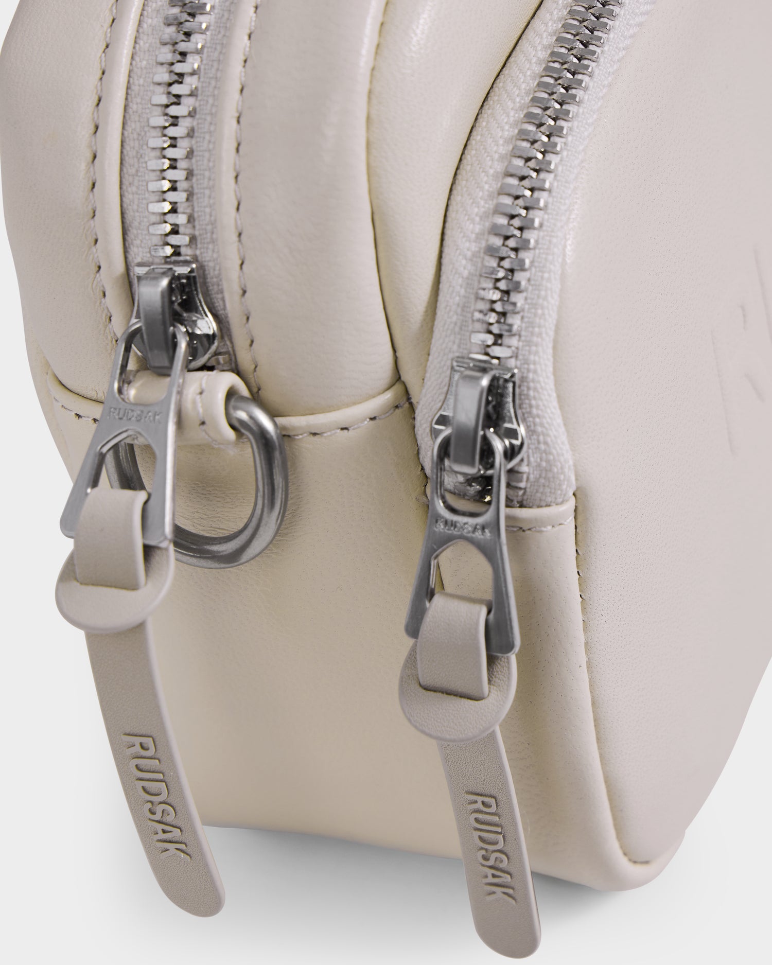 Close-up of the Rudsak ZOYA L - 8325543 Vanilla crossbody bag in beige leather, featuring two silver zippers with branded pull tabs, visible stitching, metal hardware accents, and an adjustable strap for versatile styling.