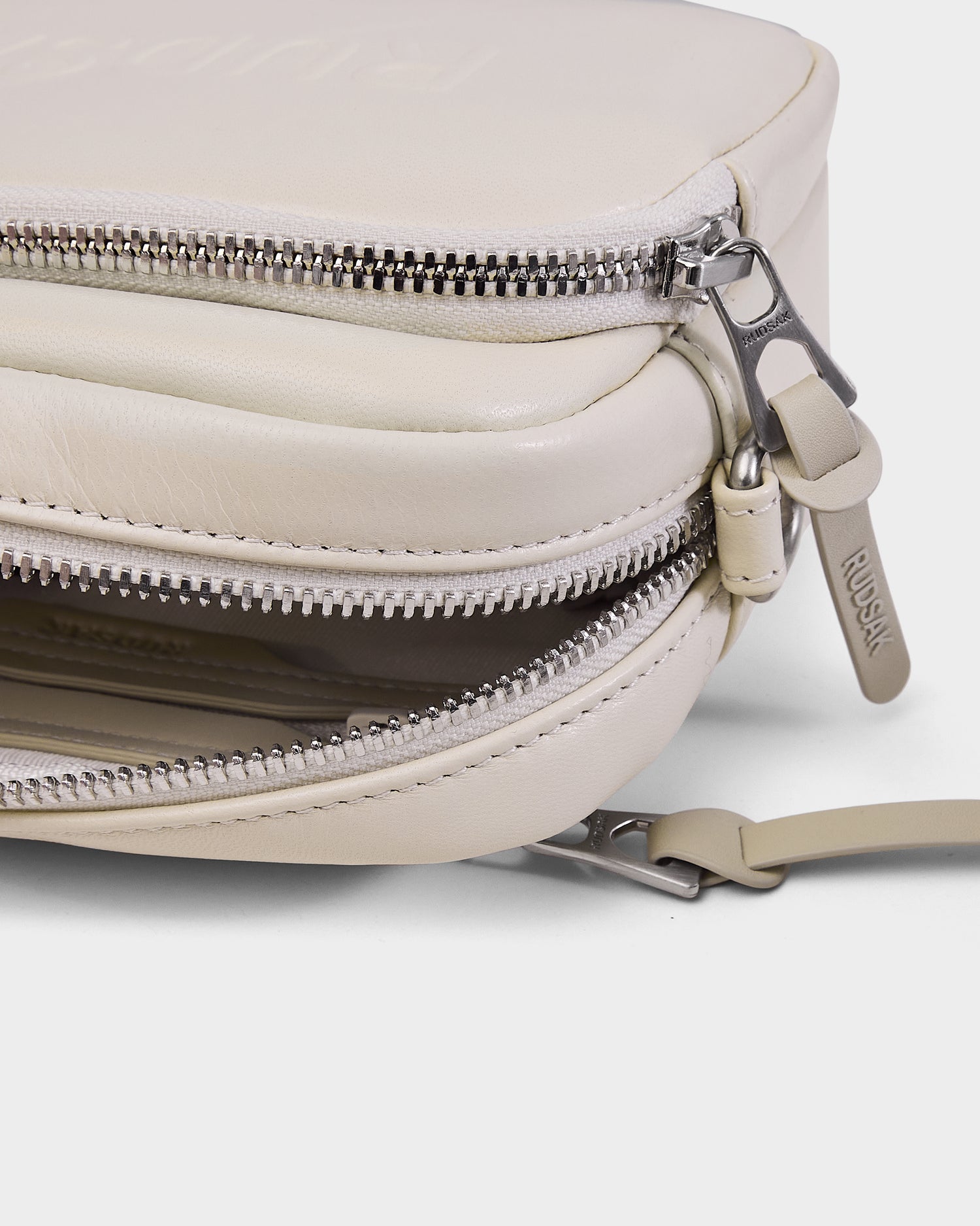 Close-up of the Rudsak ZOYA L - 8325543 Vanilla crossbody bag in cream leather, featuring two silver zippers—one slightly open—and an adjustable strap, with the Rudsak name on the zipper pull.