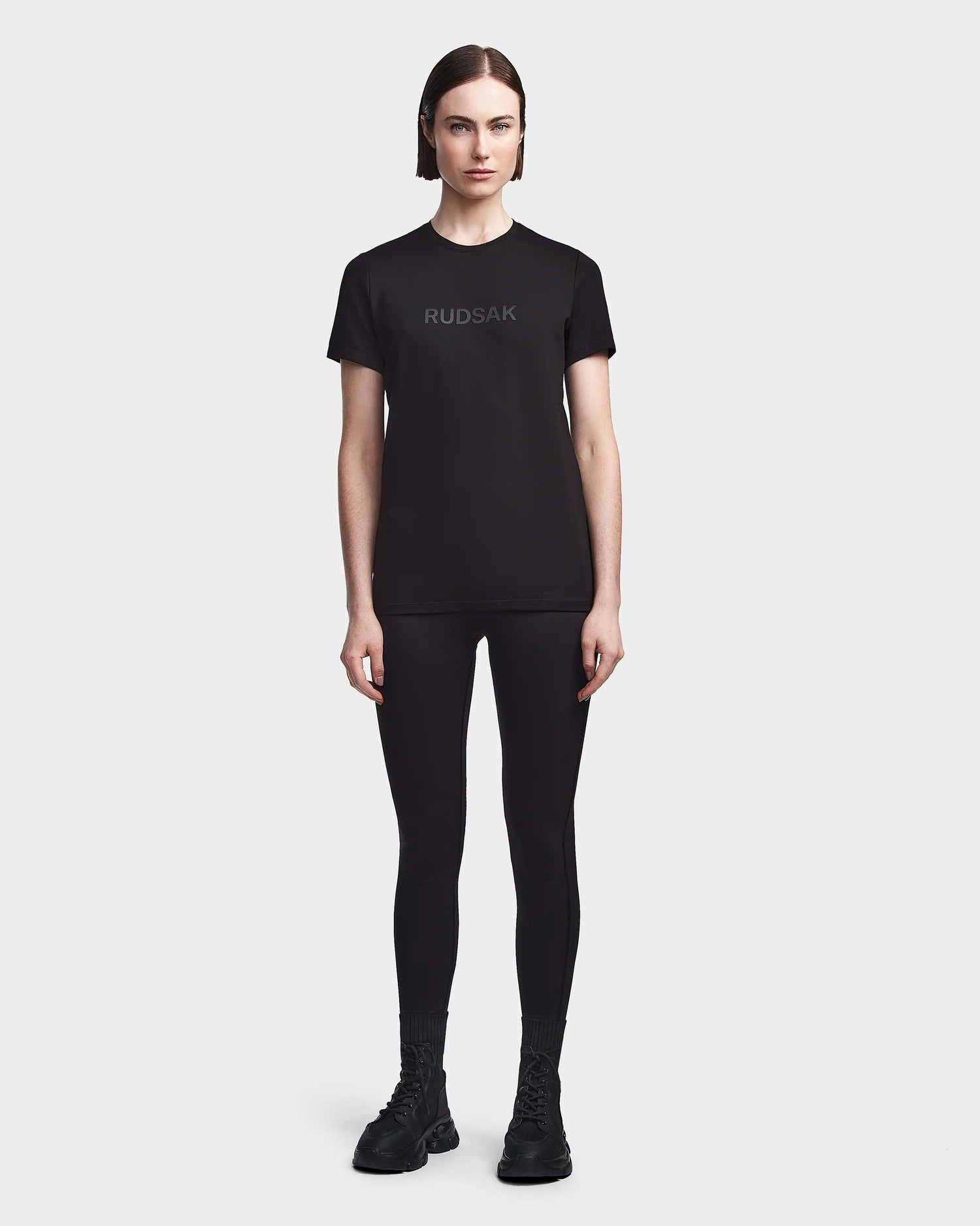 A person with straight brown hair, wearing a black RUDSAK TIFFANY - 8523048 T-shirt, black leggings, and black shoes stands facing forward against a plain white background.