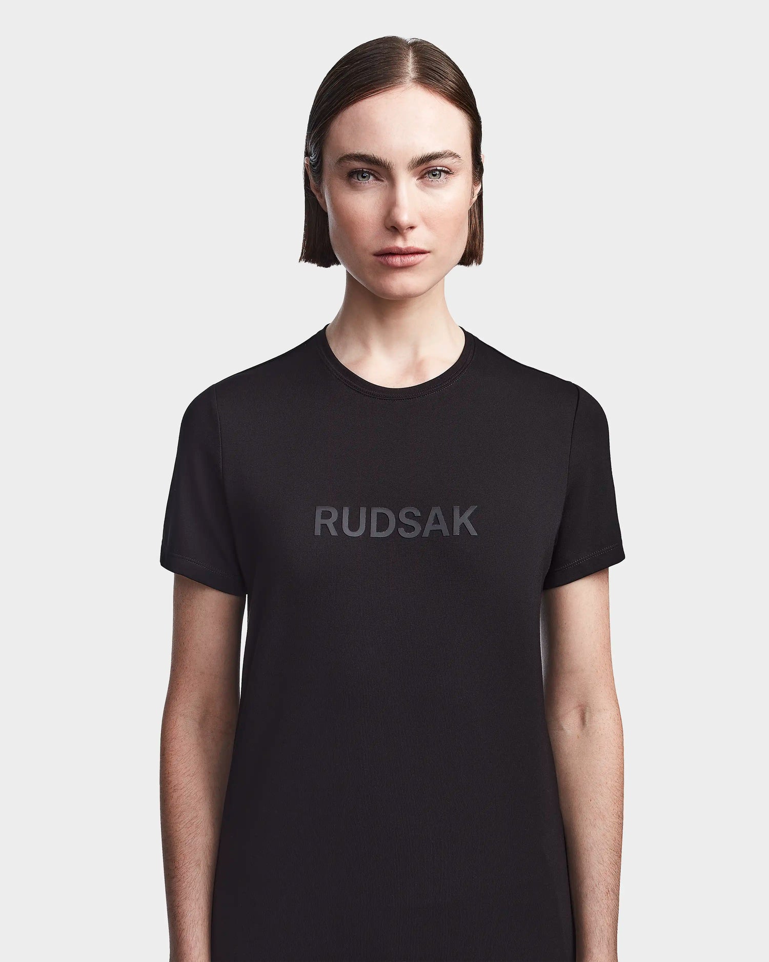 A woman with straight brown hair and blue eyes wears the RUDSAK TIFFANY - 8523048 BLACK women's T-shirt with a logo on the front, standing against a plain white background.