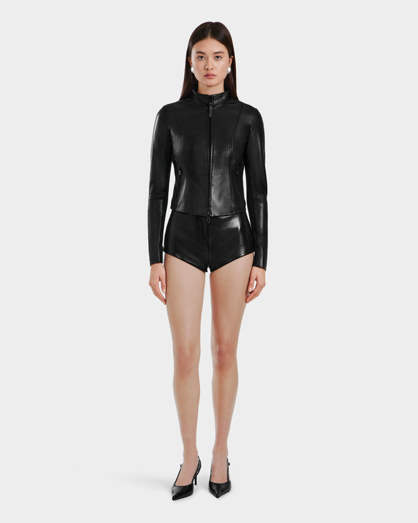 A woman stands facing forward wearing Rudsak's ABIGAIL - 8124560 Black, a fitted leather jacket with high-waisted black leather shorts and black pointed-toe heels, set against a plain white background.