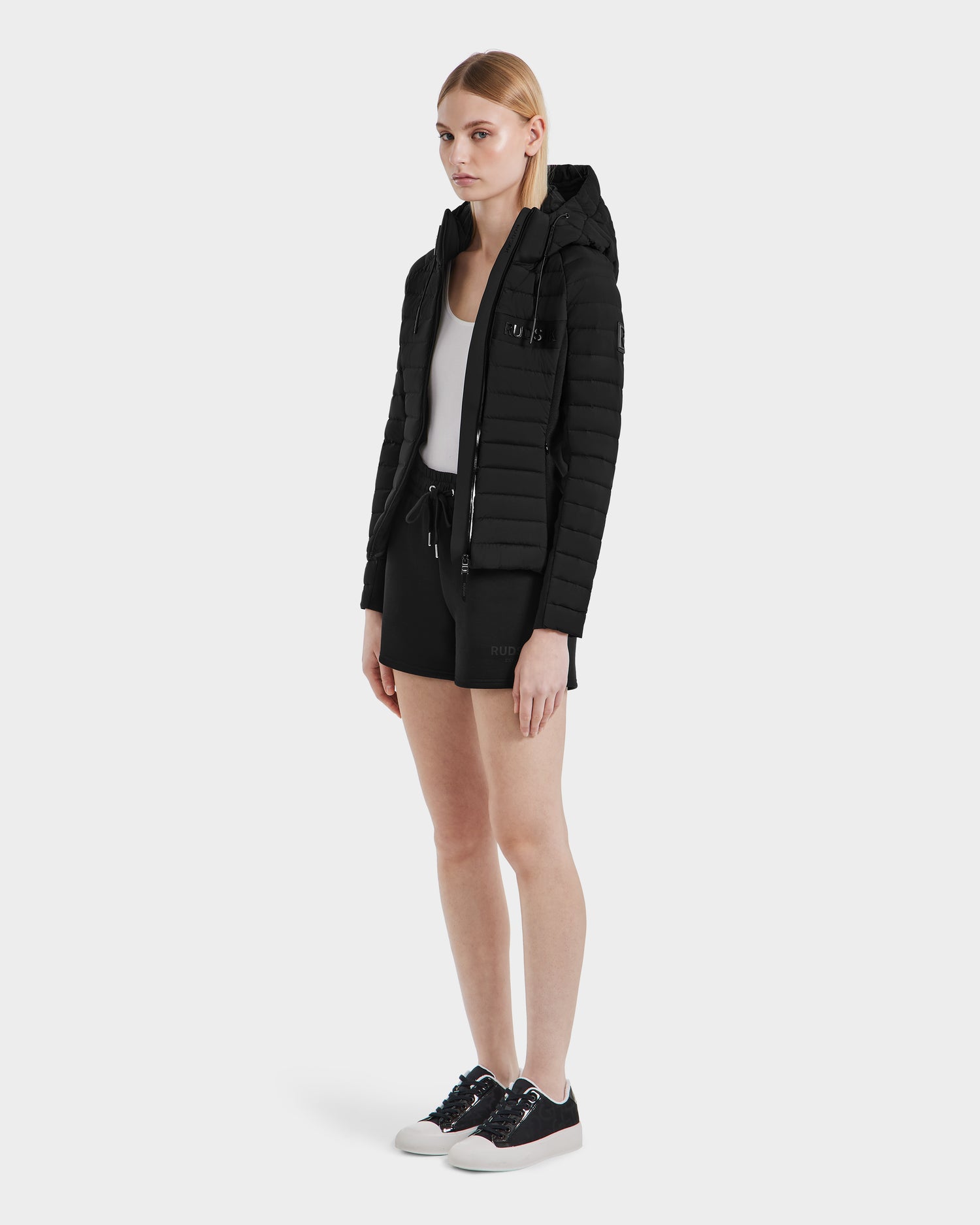 A woman faces forward against a plain white background, wearing the RUDSAK ALANA - 8123022 BLACK lightweight women's RDS-certified duck down puffer over a white top, black drawstring shorts, and black sneakers with white soles.