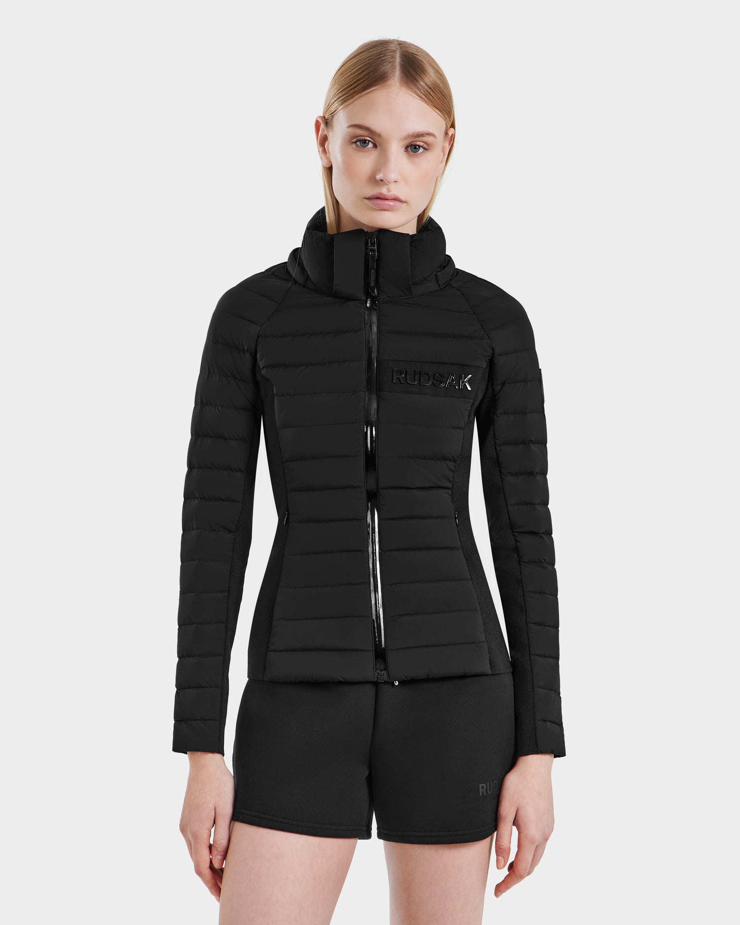 A woman with straight, blonde hair models the RUDSAK ALANA - 8123022 BLACK, an RDS-certified women's duck down jacket with a high collar, paired with black shorts against a light gray background.