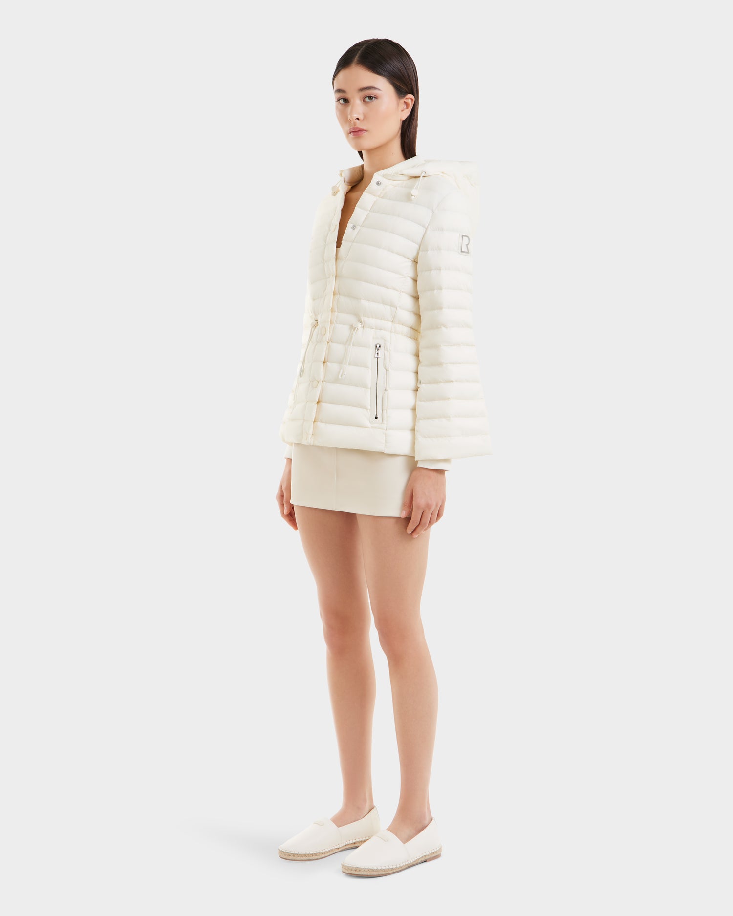 A woman poses against a plain white background in the Rudsak AMAYAH - L125015 Iceland, an RDS-certified light down puffer jacket with a hood, styled with a cream mini skirt and slip-on shoes for a chic, feminine look.