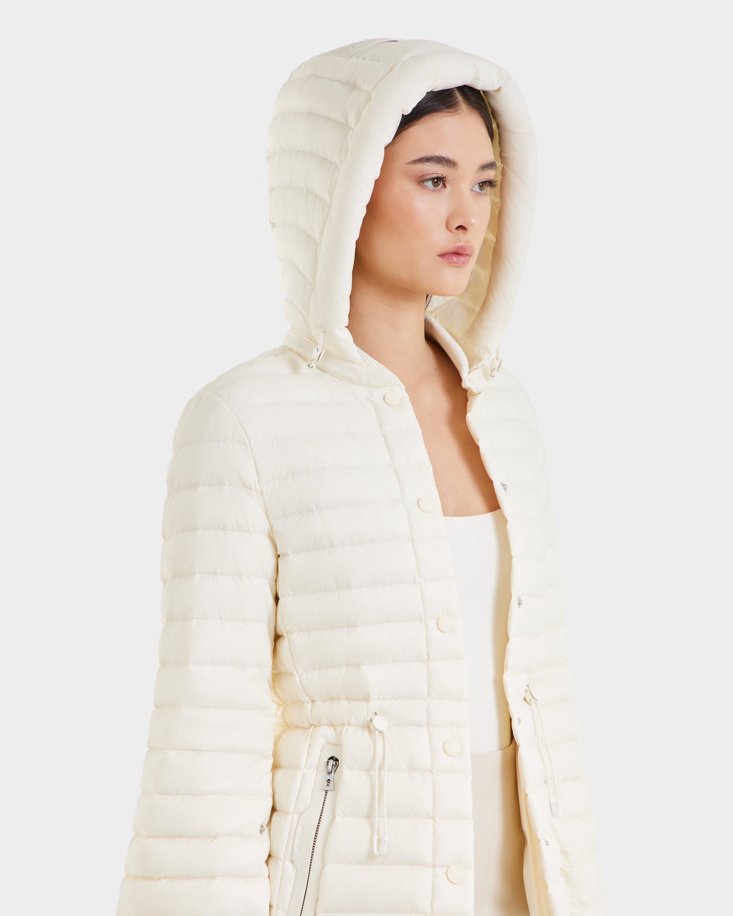 A woman wears the Rudsak AMAYAH - L125015 Iceland off-white quilted lightweight down jacket with a feminine silhouette, hood, front zipper, and snap buttons, standing against a plain white background.