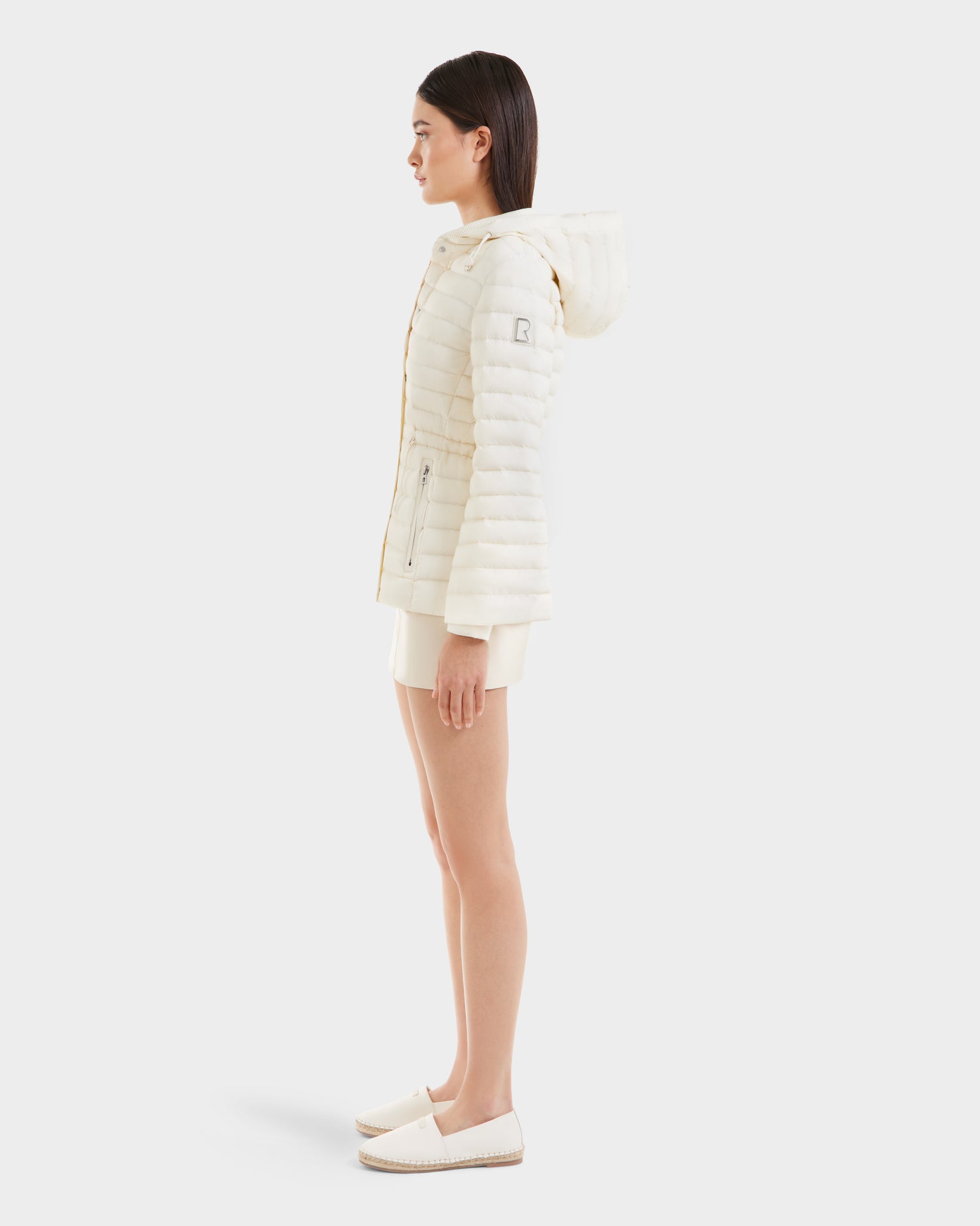 A woman in profile wears the Rudsak AMAYAH - L125015 Iceland off-white, hooded, lightweight down jacket with a feminine silhouette, paired with matching shorts and white slip-on shoes against a plain white background.