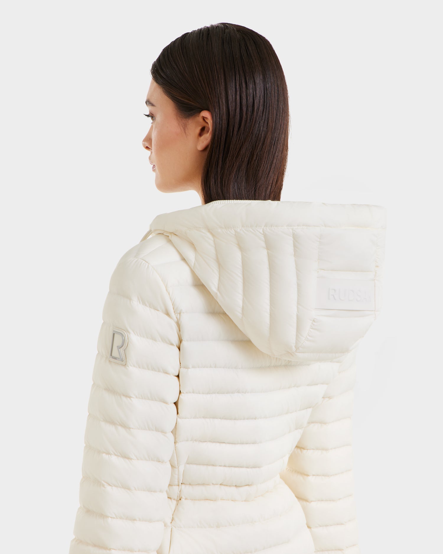 A woman with straight brown hair wears the Rudsak AMAYAH - L125015 Iceland cream lightweight down jacket, featuring a feminine silhouette, sleeve patch, and RUDSAK text on the hood. Shown from back and side against a white background.