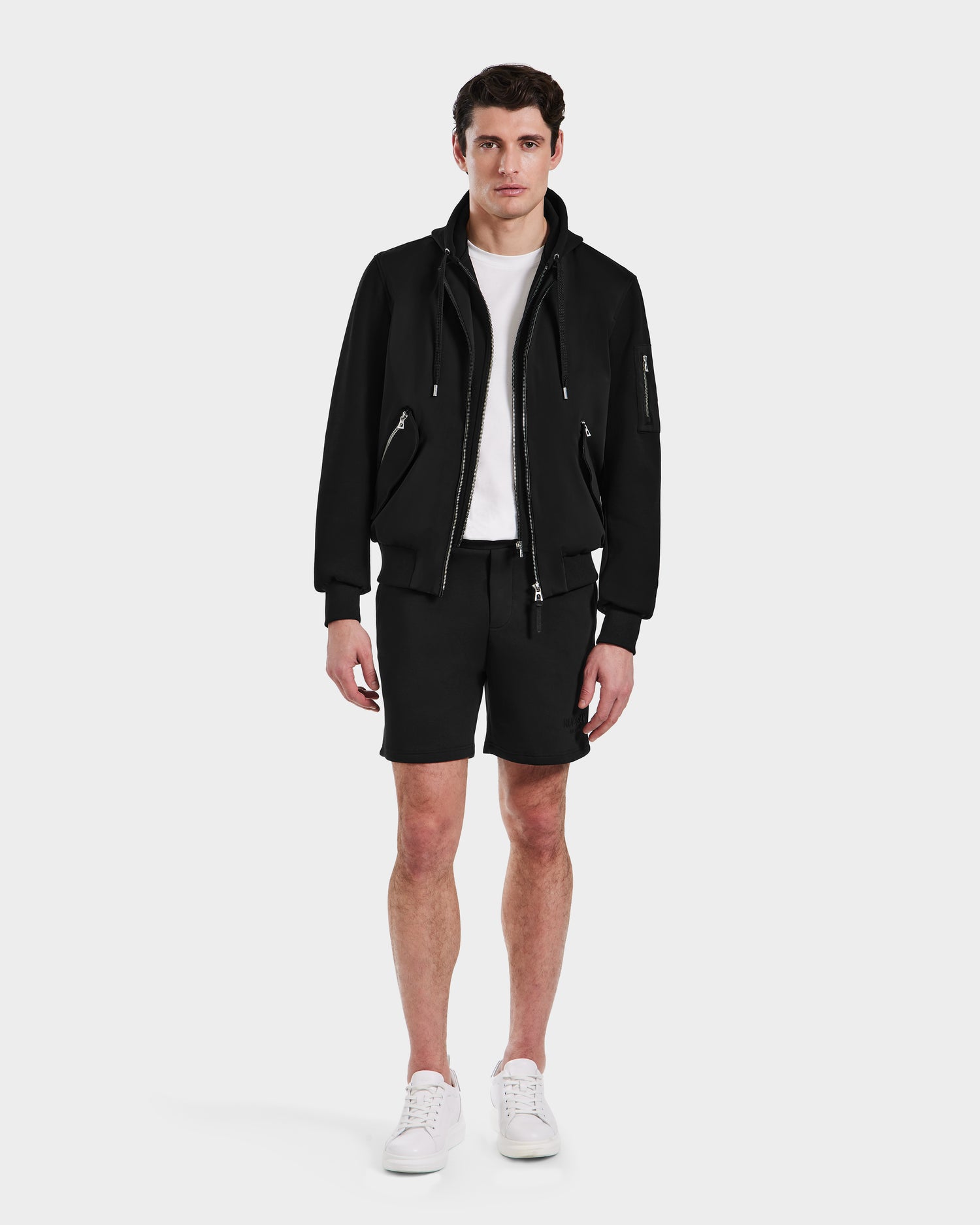 A man faces forward in a Rudsak ANDRE - 6124020 Black men's lightweight bomber jacket with removable hooded bib, white t-shirt, black shorts, and white sneakers against a plain white background.