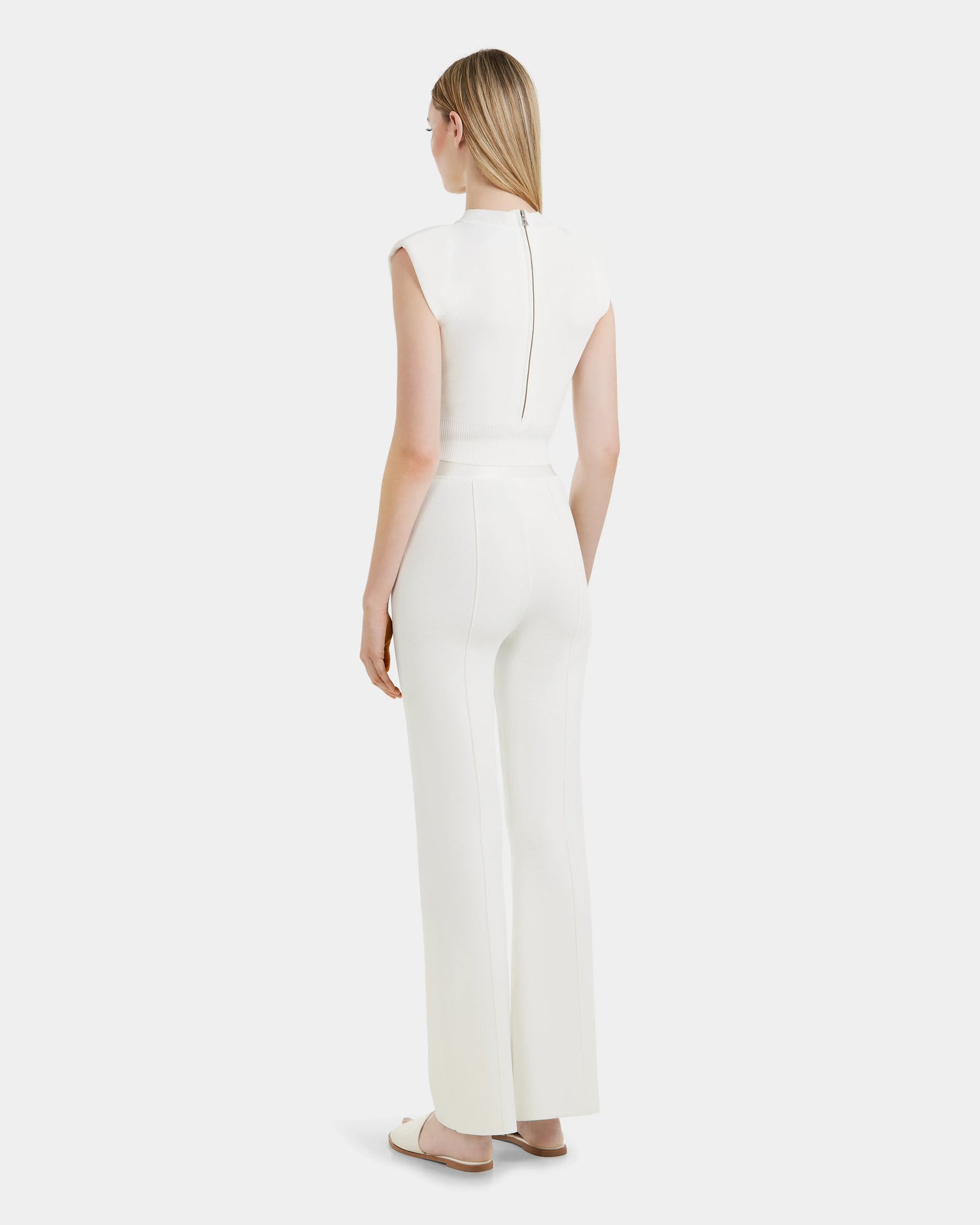 A woman with straight blonde hair faces away, wearing the ANGIE - 8524008 Cotton White sleeveless top and high-waisted wide-leg pants from Rudsak, paired with white slide sandals against a light background.