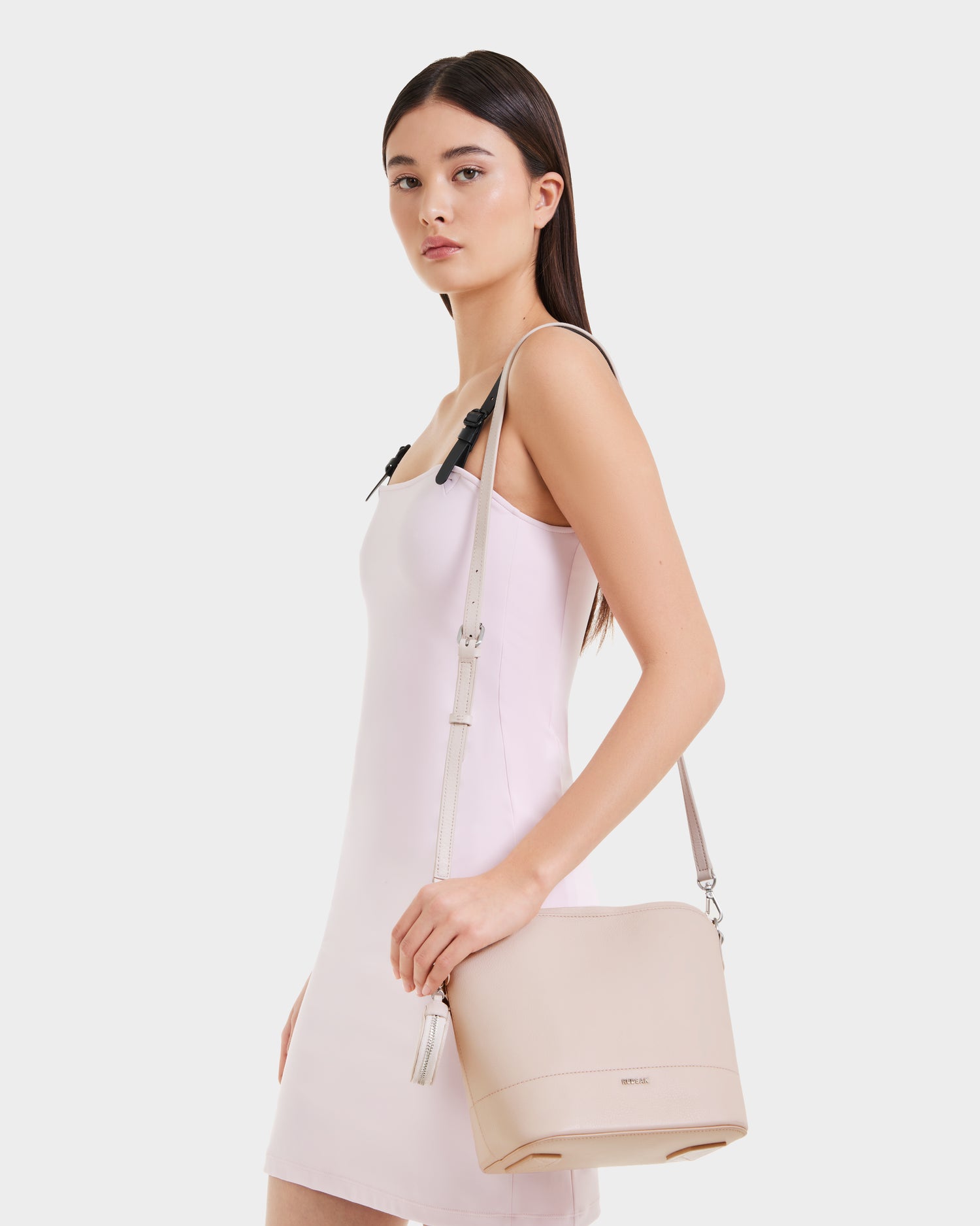 A woman in a pale pink sleeveless dress poses sideways with the Rudsak ANIA SM - 8325009 Soft Pink compact hobo tote, featuring an adjustable crossbody strap and tassel detail, against a plain white background.