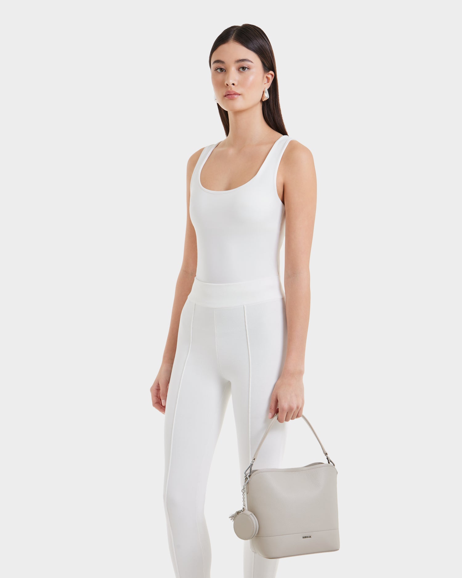 A woman with long dark hair, dressed in a fitted white tank top and white pants, poses against a plain white background holding the Rudsak ANIA SM - 8325009 Vanilla pebble leather hobo bag with attached small round pouch.