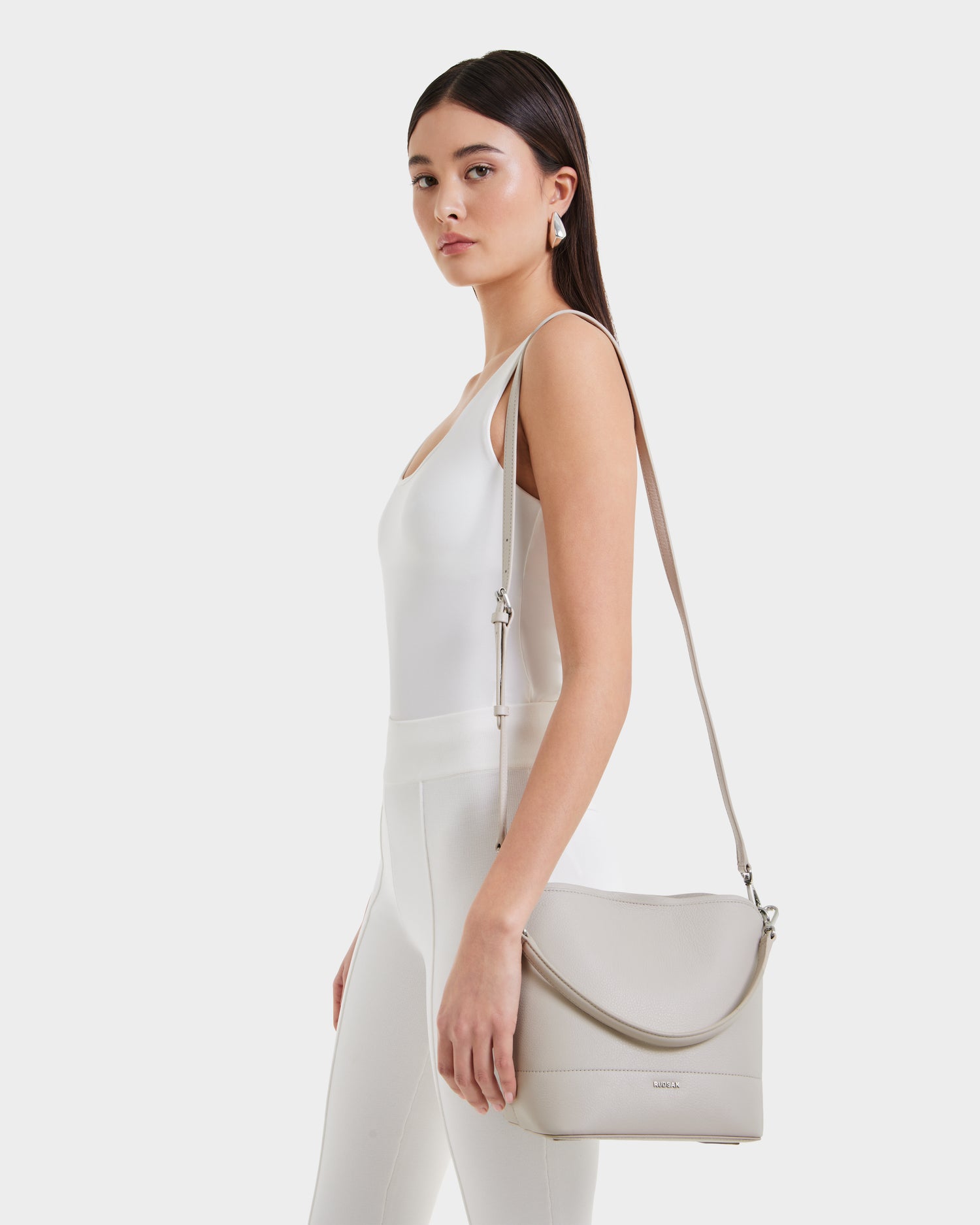 A woman in a white sleeveless top and pants stands sideways, showcasing the Rudsak ANIA SM - 8325009 Vanilla pebble leather hobo bag with a crossbody strap. The background is plain white.