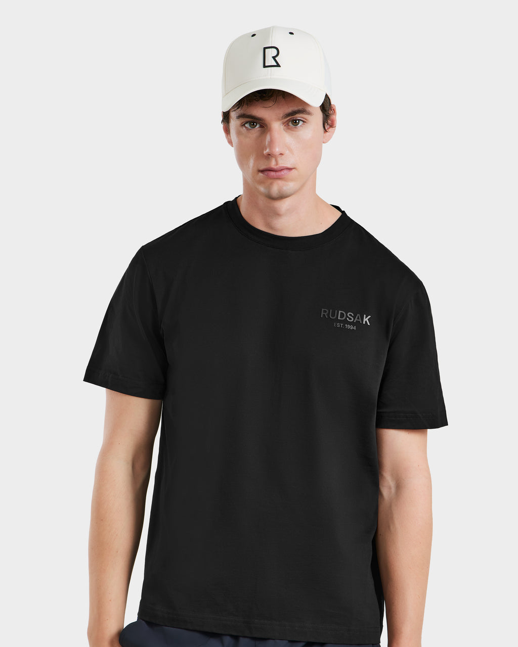 A young man wears the Rudsak ARSENE - 9422504 Iceland unisex baseball cap and a black T-shirt with RUDSAK EST 1994 on the chest, standing against a plain light background and looking at the camera with a neutral expression.