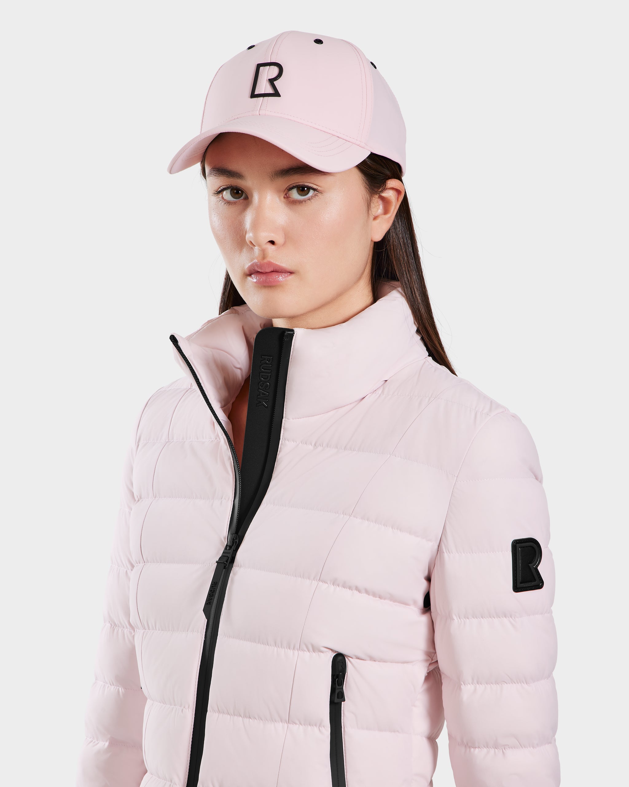 A woman with straight brown hair and a neutral expression wears the Rudsak ARSENE - 9422504 light pink quilted jacket with black zippers and a matching unisex baseball cap, both featuring a black R logo.