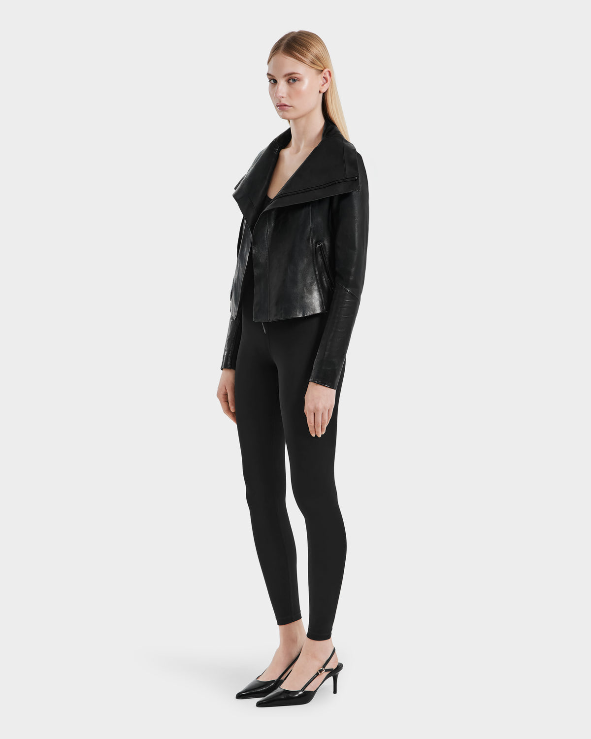 A woman stands against a plain background wearing the Rudsak AVRIL - 8124561 Black womens leather jacket, black leggings, and heels, with her hair down and hands by her sides.