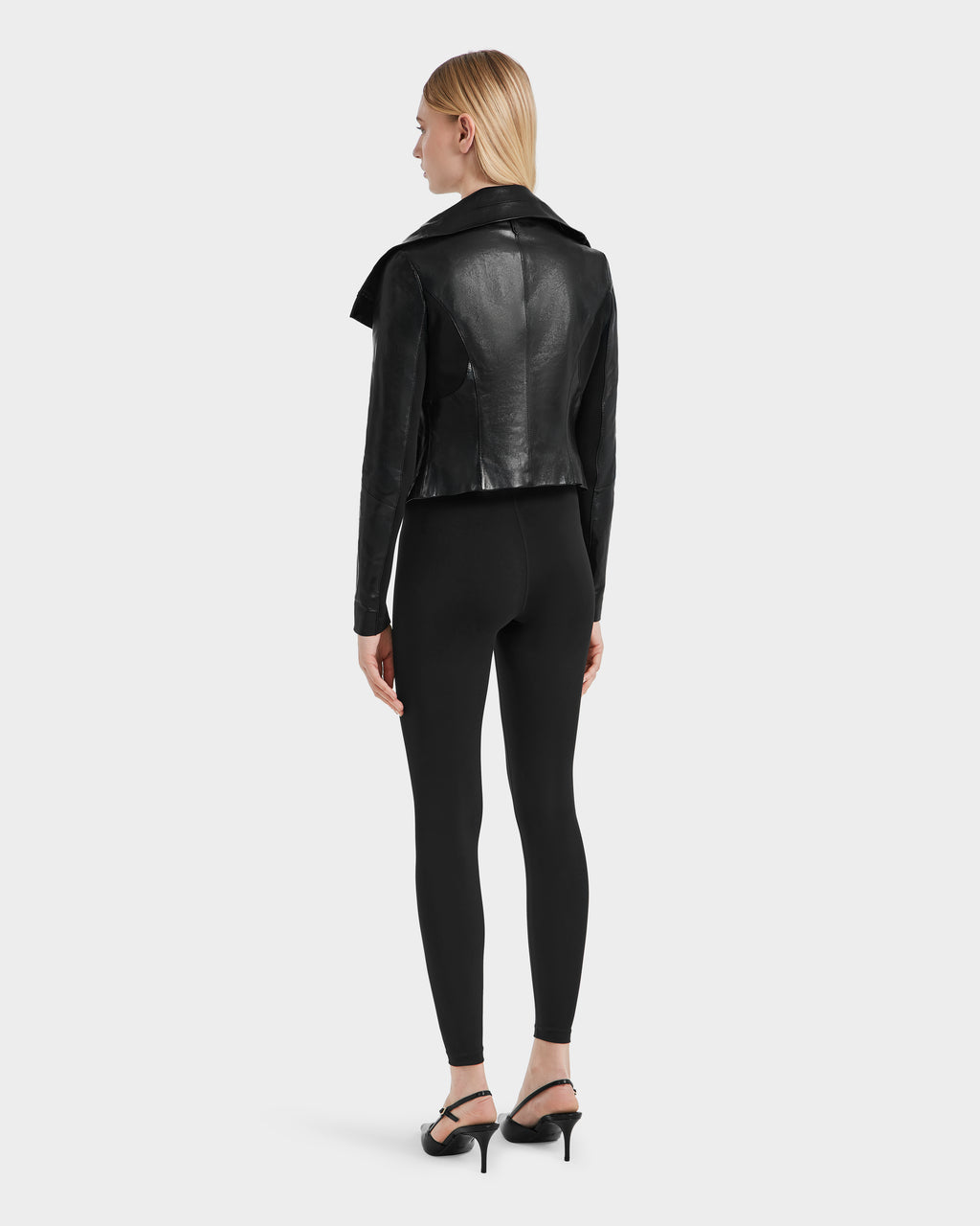 A woman with straight blonde hair stands facing away, wearing the Rudsak AVRIL - 8124561 Black womens leather jacket, paired with black leggings and heeled sandals against a plain light background.