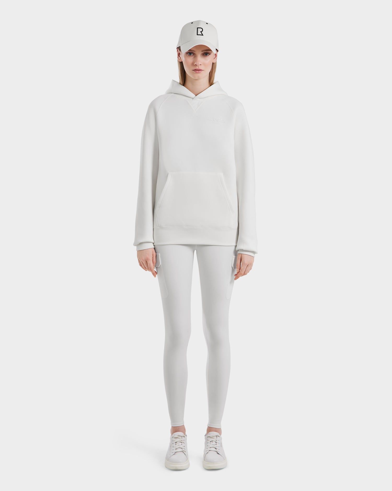 A person faces forward in an all-white outfit, featuring the Rudsak BARRY - 9524011 Iceland W hoodie, leggings, sneakers, and a baseball cap with a black R logo. The background is plain white.