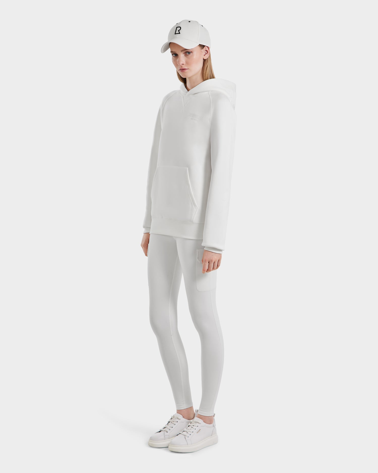 A person stands against a white background wearing the Rudsak BARRY - 9524011 Iceland W oversized unisex hoodie, paired with white leggings, sneakers, and a baseball cap for a casual, monochromatic look with hands by their sides.