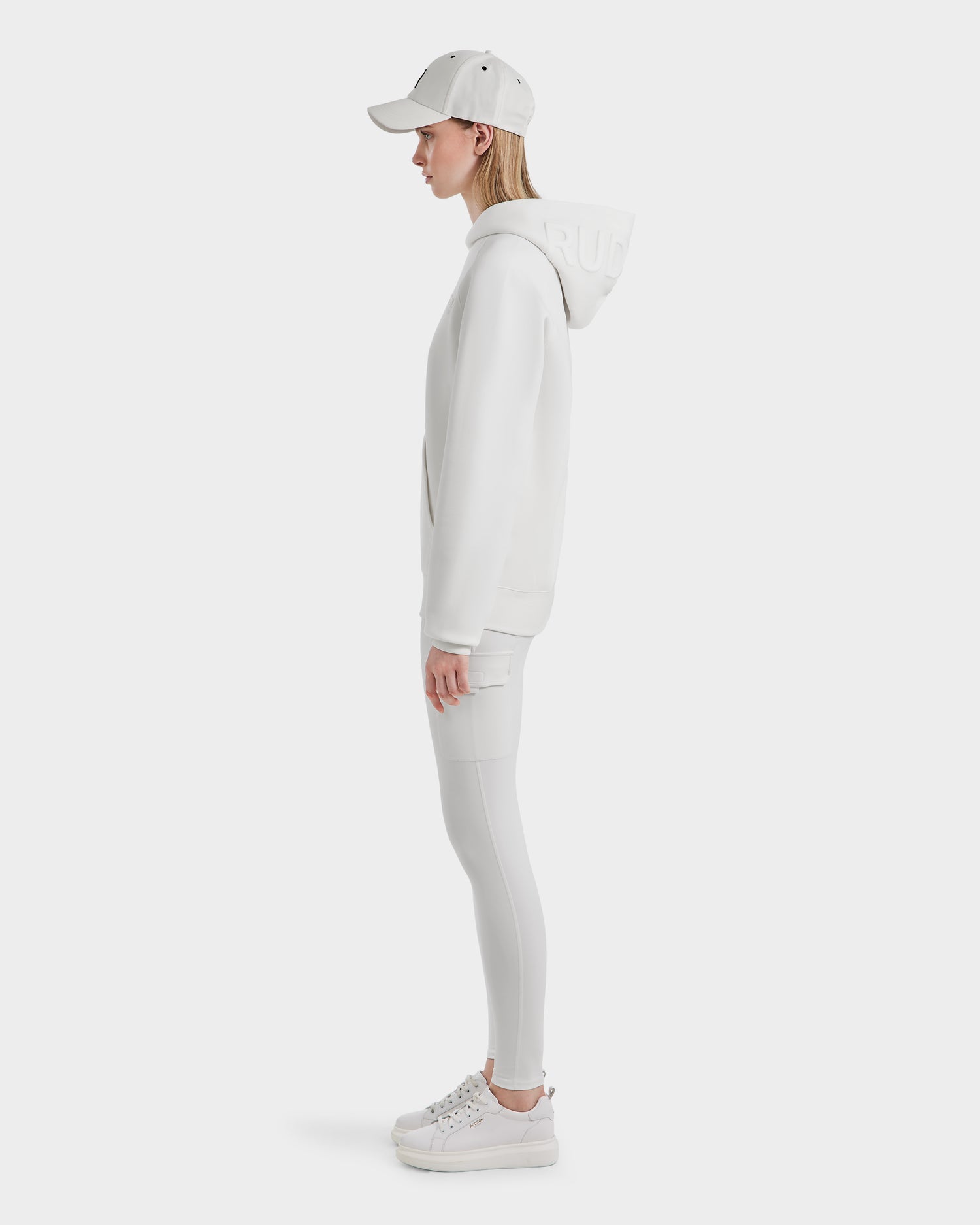 A person stands in profile, wearing the oversized Rudsak BARRY - 9524011 Iceland W hoodie with the hood up, paired with white leggings, sneakers, and a baseball cap against a plain white background.