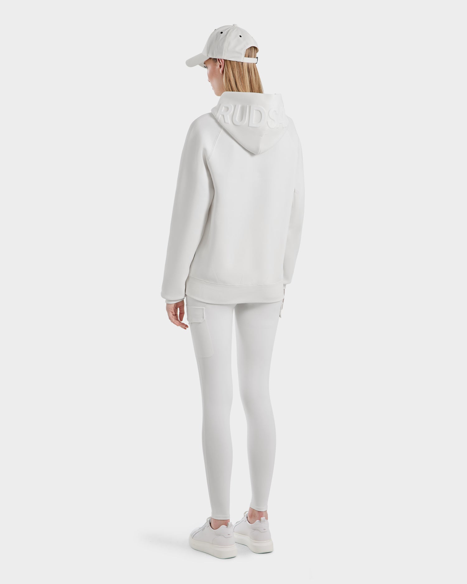 A person faces away, wearing the Rudsak BARRY - 9524011 Iceland W hoodie with RUDS embossed on the hood, paired with white leggings, sneakers, and a baseball cap against a plain white background.