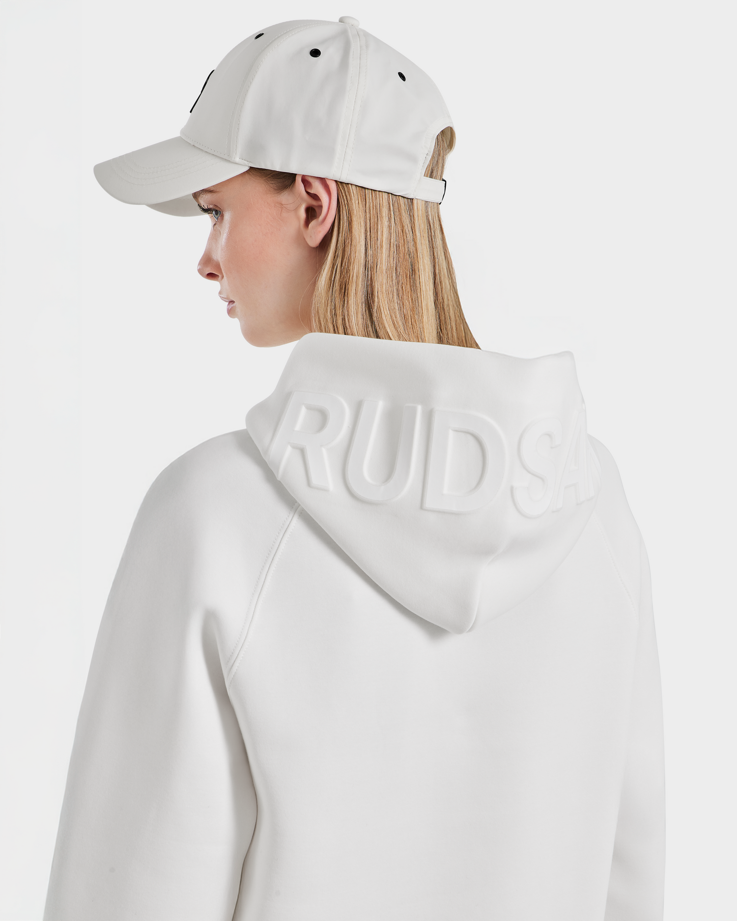 A person wearing the oversized Rudsak BARRY - 9524011 Iceland W white hoodie with embossed branding on the hood and a white baseball cap, shown from the back and in profile against a plain white background.