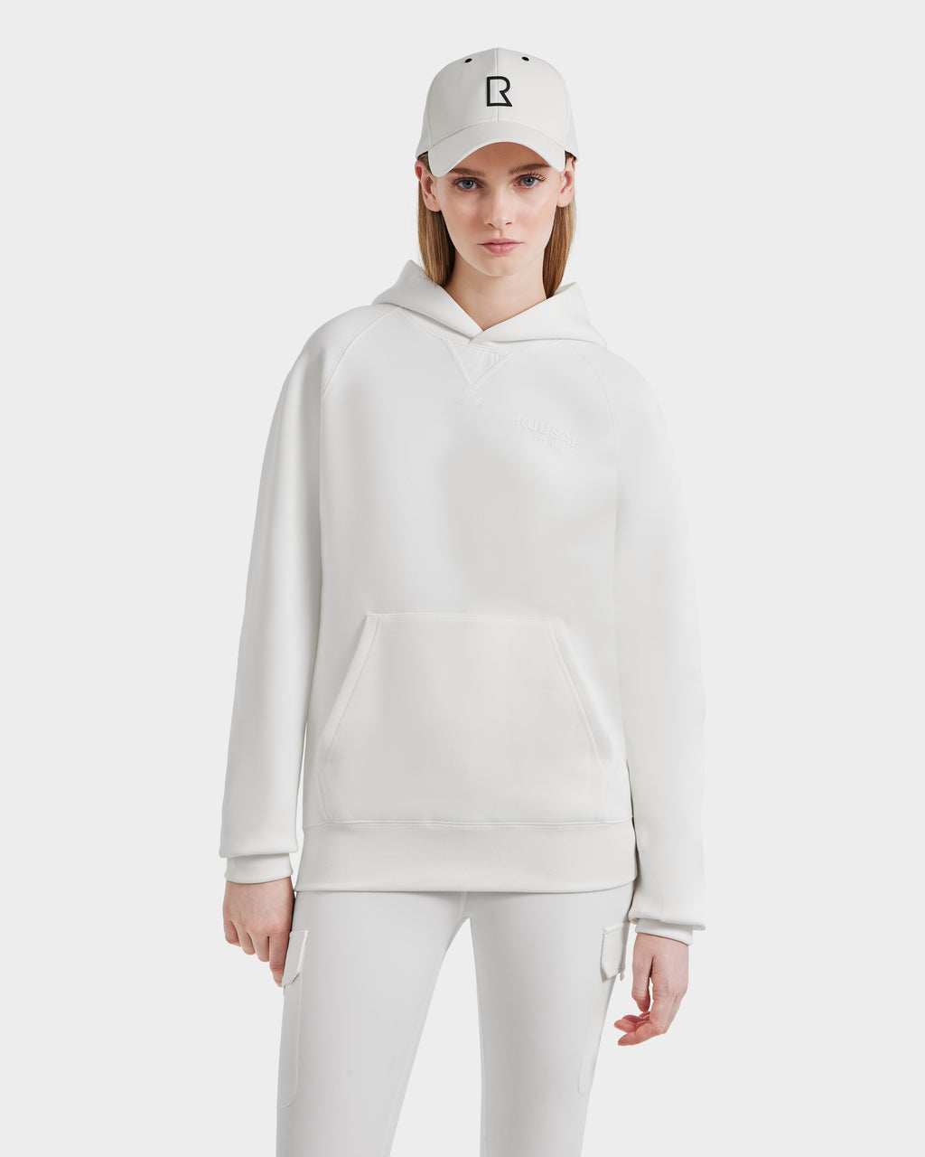 A woman faces forward against a plain white background, wearing the all-white Rudsak BARRY - 9524011 Iceland W hoodie, fitted pants, and a baseball cap with a black R logo.