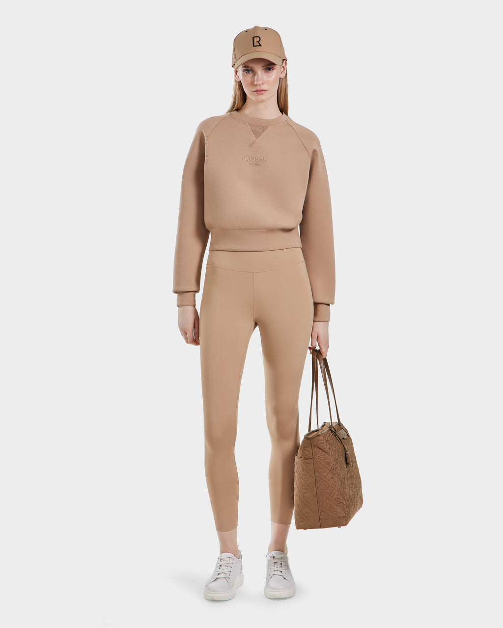 A woman stands against a white background wearing the Rudsak BETSY - 8525087 Almond crewneck sweater with matching leggings, white sneakers, a tan cap, and holding a tan duffle bag in her left hand.