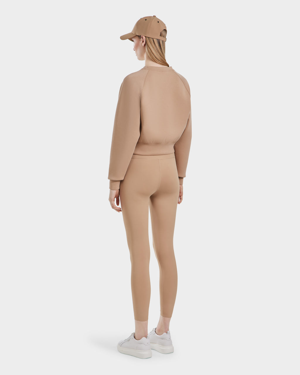 A woman stands facing away, styled in the Rudsak BETSY - 8525087 Almond sweater and matching leggings with a beige cap and white sneakers, all set against a plain white background.