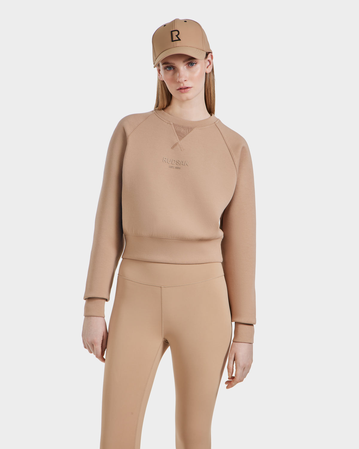A woman in the Rudsak BETSY - 8525087 Almond premium cotton blend crewneck sweater, matching leggings, and a baseball cap stands against a white background, looking at the camera.