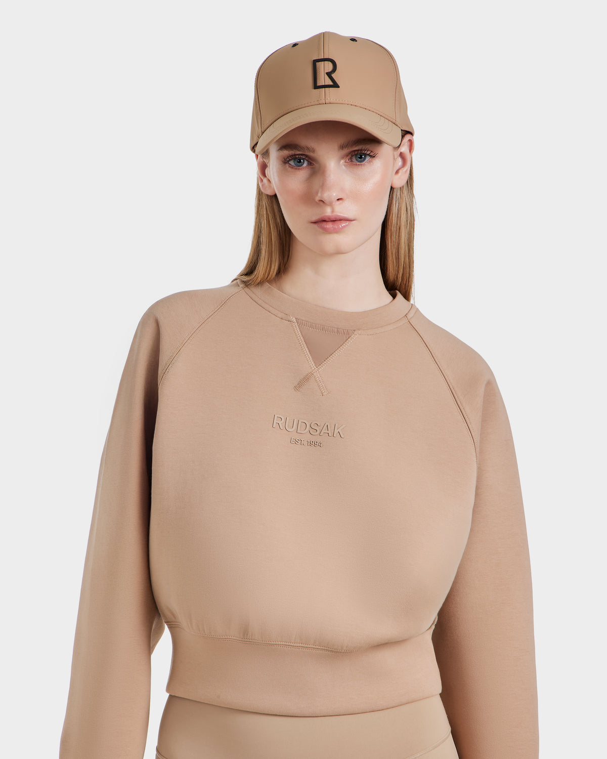 A woman in the Rudsak BETSY - 8525087 Almond crewneck sweater with RUDSAK embossed on the front and a matching beige cap with a black R logo stands against a plain white background.