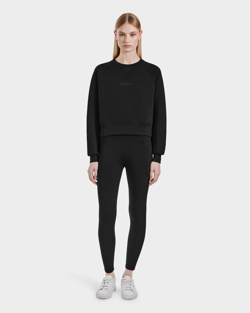 A woman with straight blonde hair faces forward, wearing the Rudsak BETSY - 8525087 Black women’s crop sweater, paired with black leggings and white sneakers, set against a plain light background.