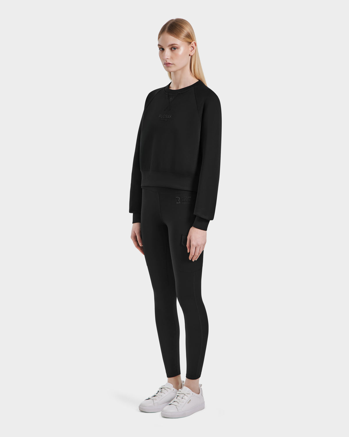 A woman models the Rudsak BETSY - 8525087 Black crop sweater against a white background, paired with black leggings and white sneakers. She stands slightly turned left, arms relaxed at her sides.