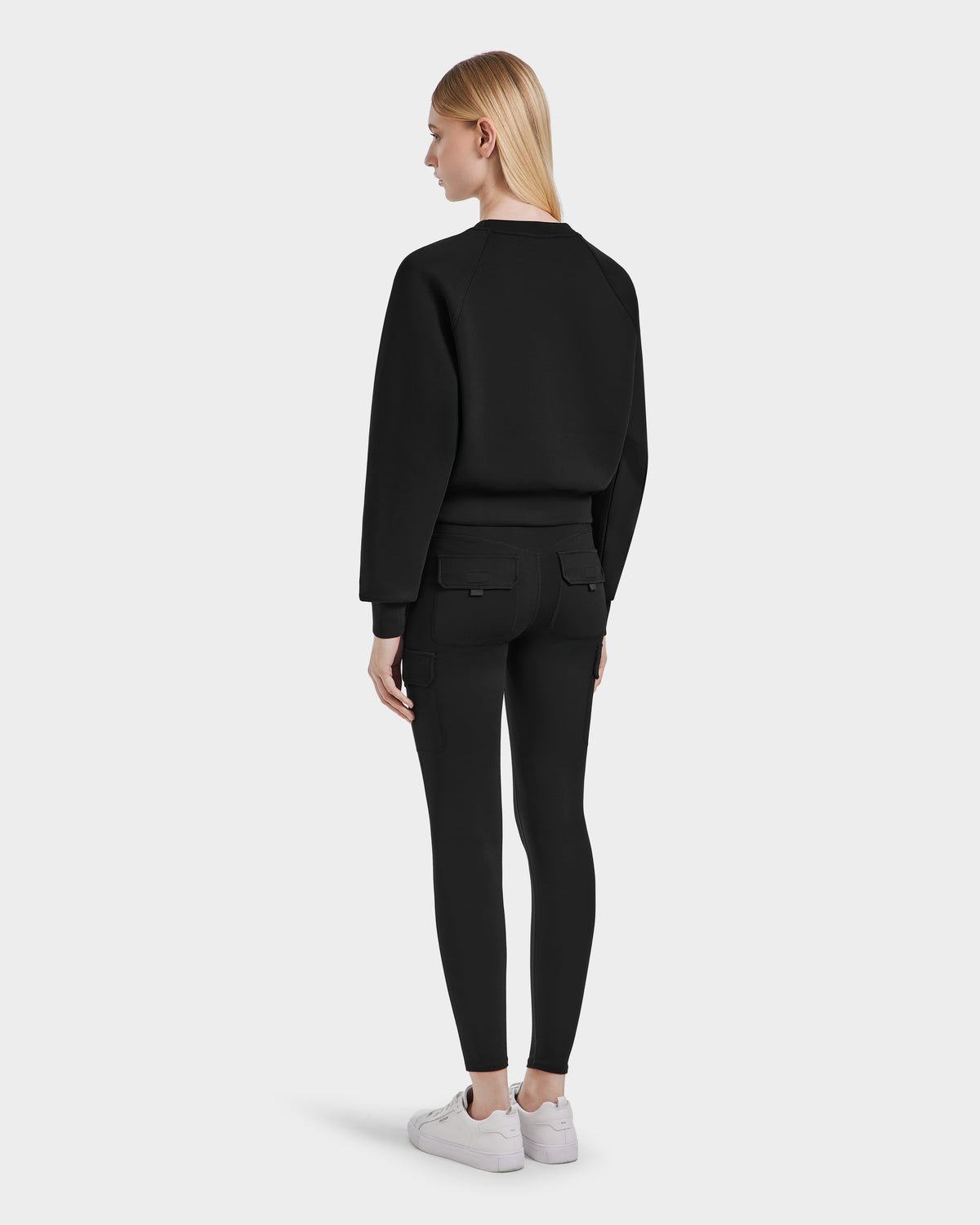 A woman with straight blonde hair, facing away, wears the Rudsak BETSY - 8525087 Black premium cotton blend womens crop sweater, paired with black fitted pants and white sneakers against a plain light gray background.